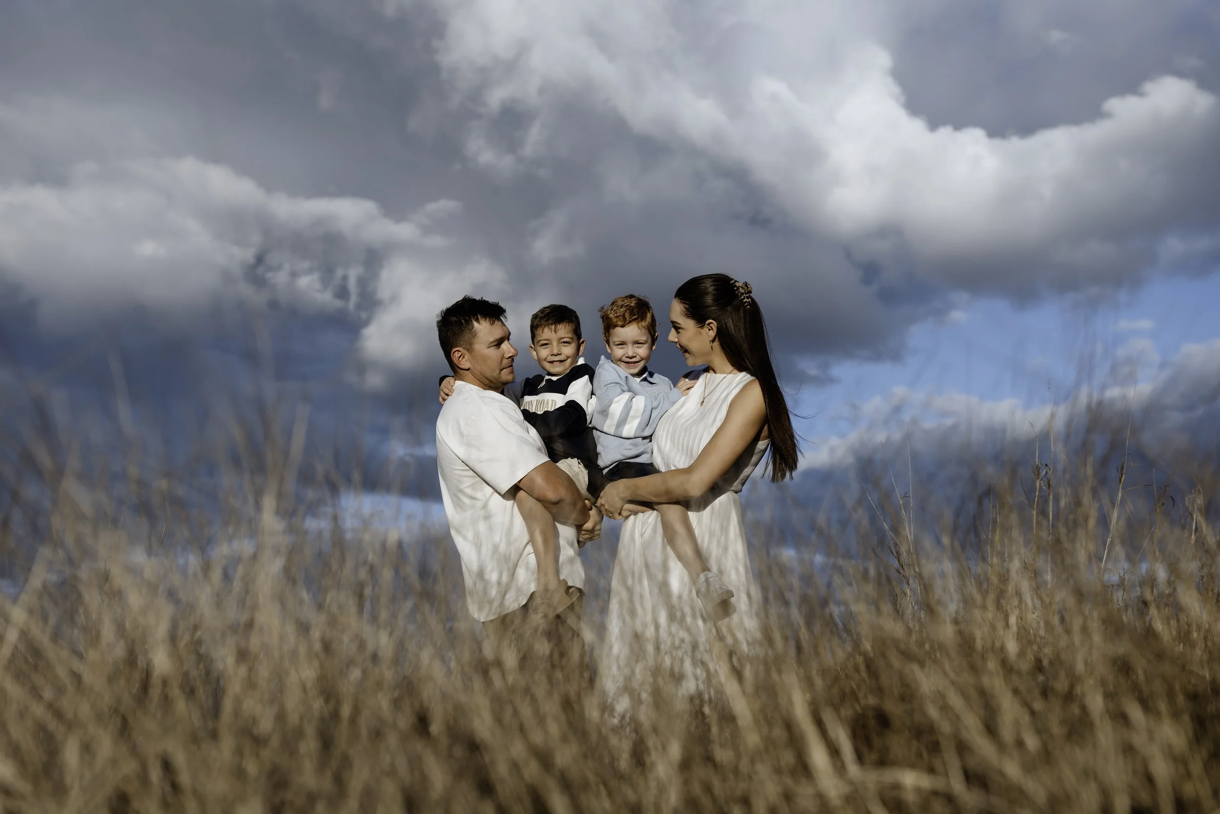 A family of four with two young boys standing in a grassy field under a cloudy sky. The father and mother are smiling and holding the boys, creating a warm family scene.