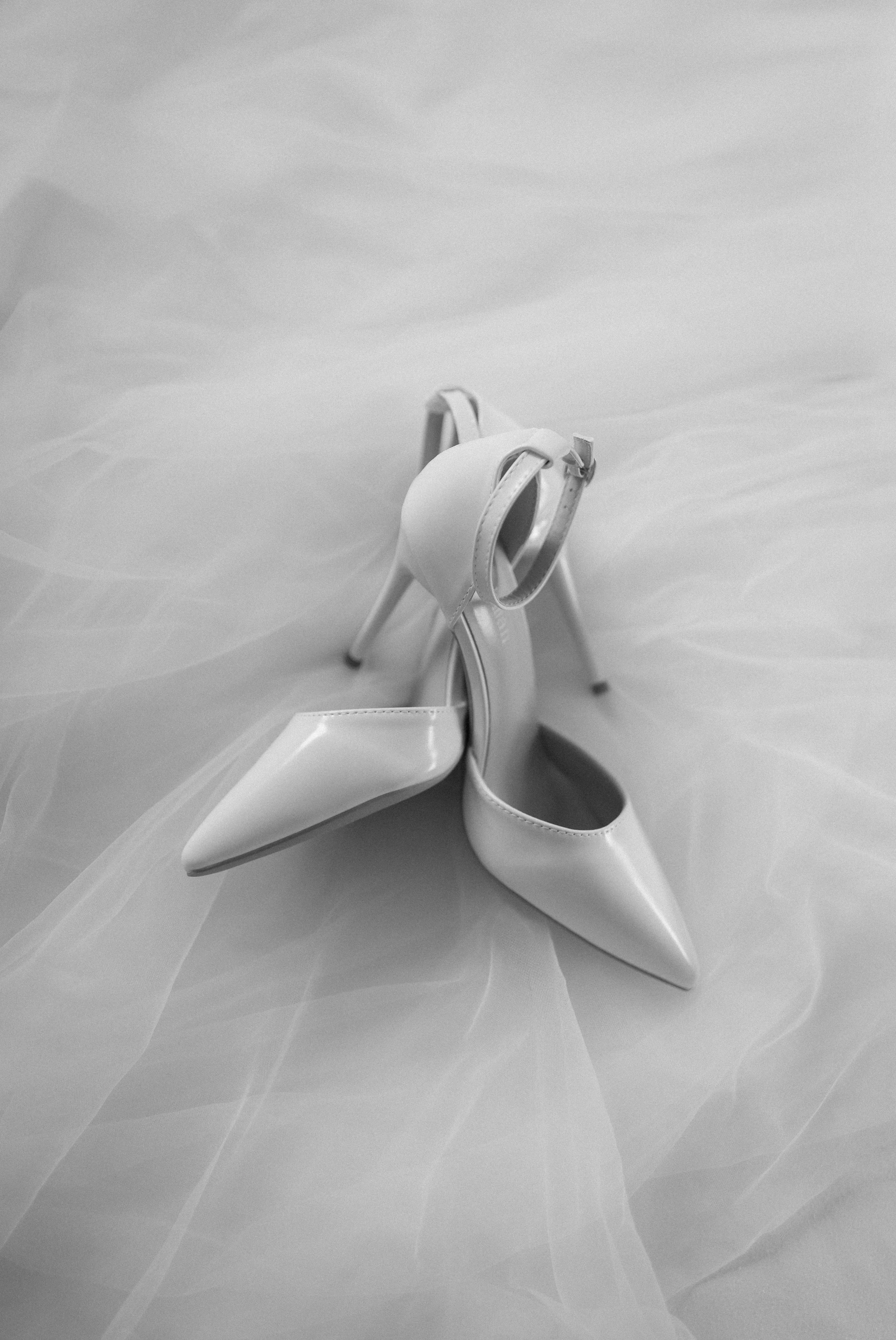 A black-and-white image of a pair of white high-heeled shoes with ankle straps, placed on a sheer, ruffled fabric background.