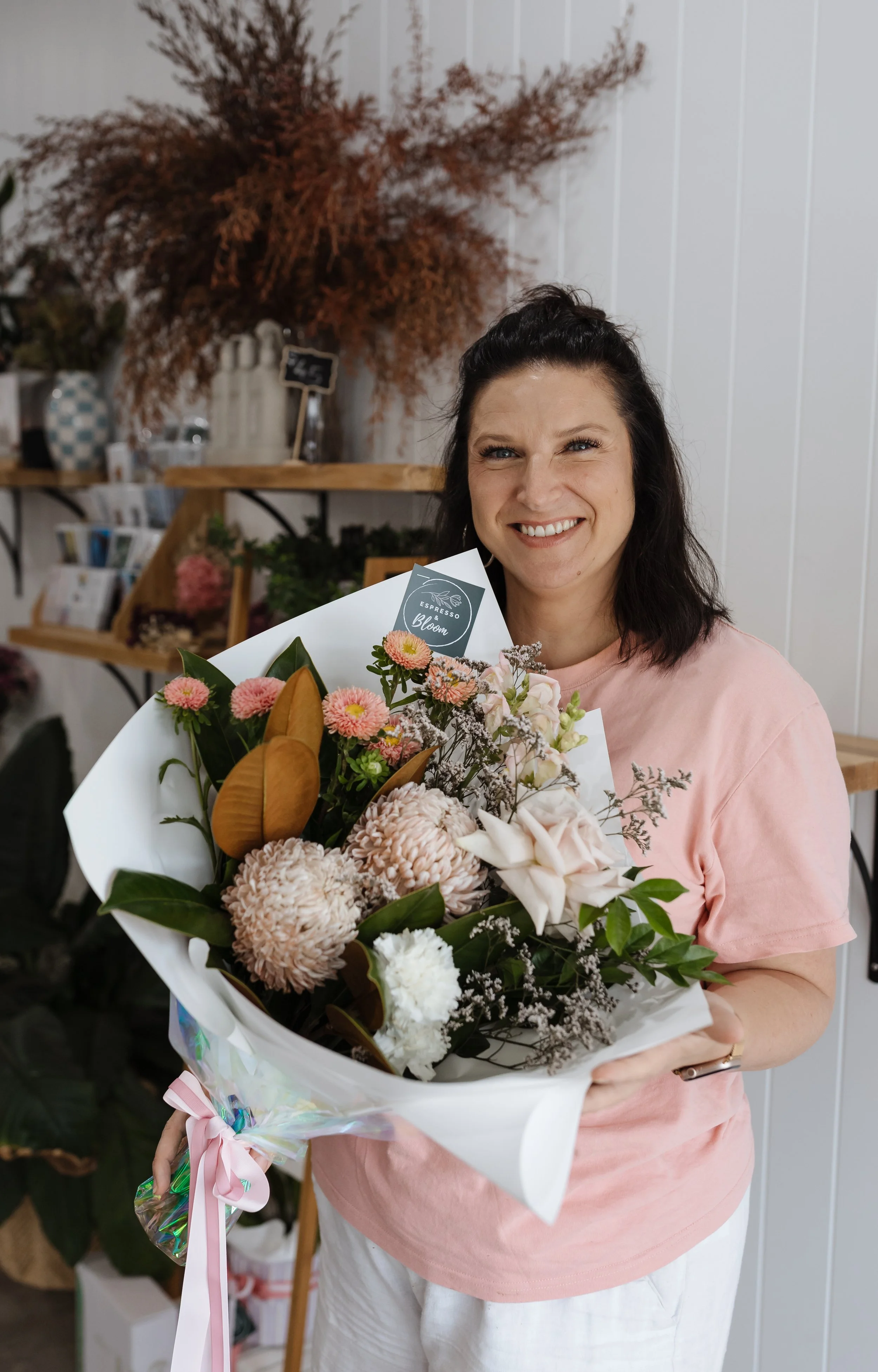 A woman with dark hair styled in a half-updo, smiling and holding a large bouquet of mixed flowers including pink, white, and peach blooms, in a flower shop or boutique setting.