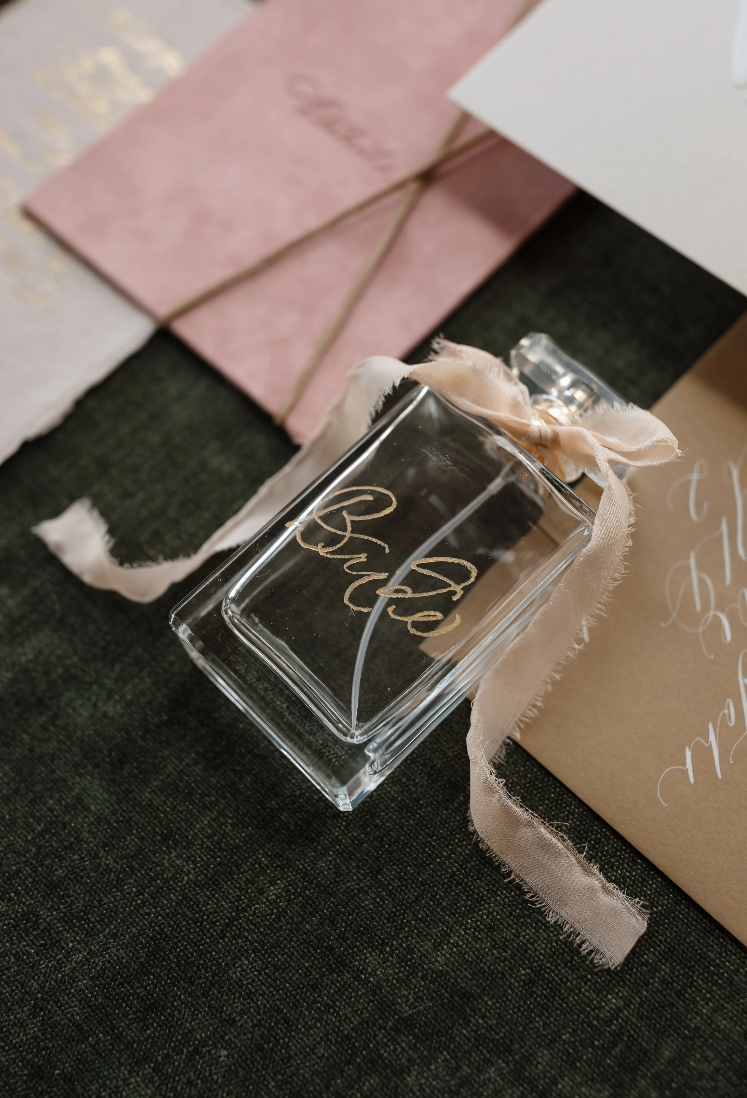 A clear glass perfume bottle with a fabric ribbon tied around its neck, placed on a dark textured surface. In the background, there are pink and white gift boxes, with the pink box tied with a brown string and the white box partially visible.