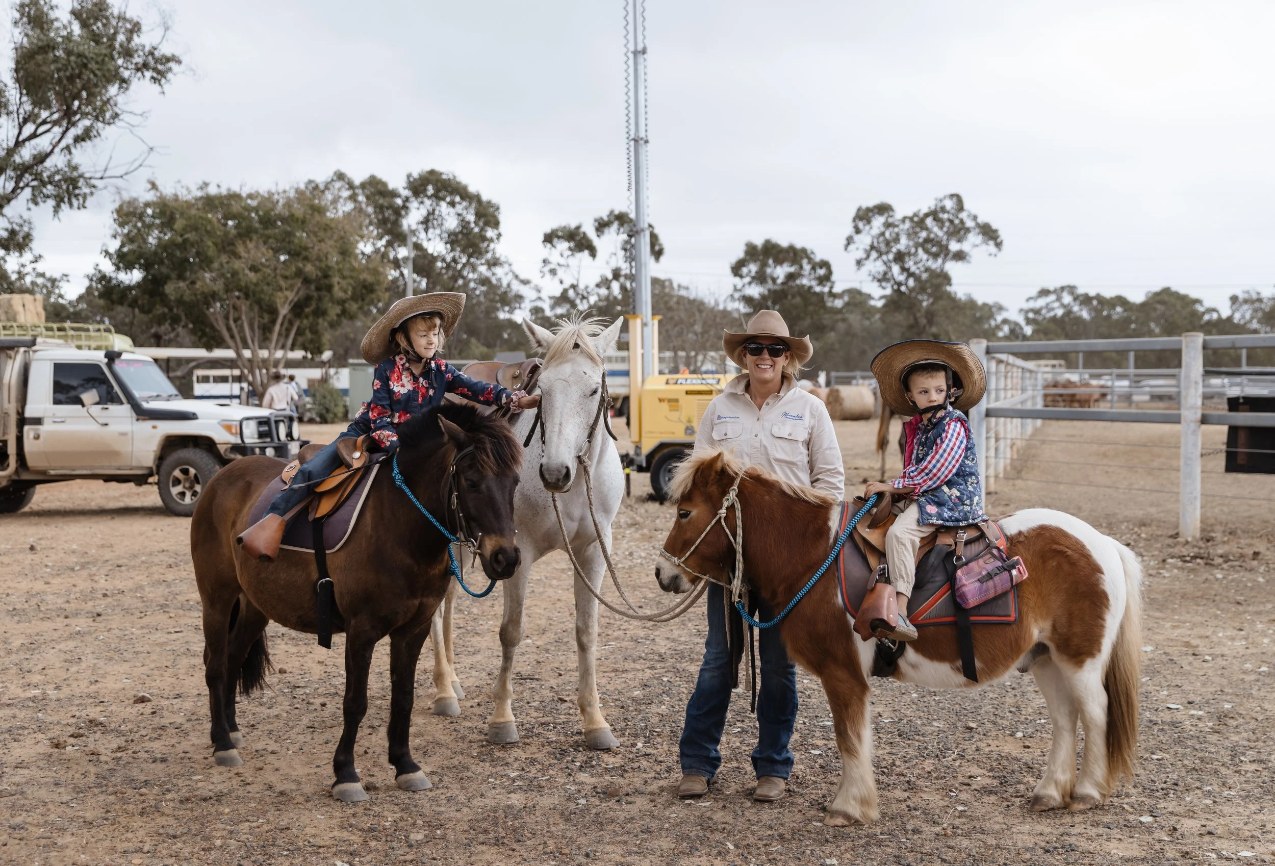 Three children and an adult riding and leading horses at an outdoor riding area. The children are wearing hats and plaid shirts. The adult is wearing sunglasses and a cowboy hat. There are trees, vehicles, and a metal fence in the background.