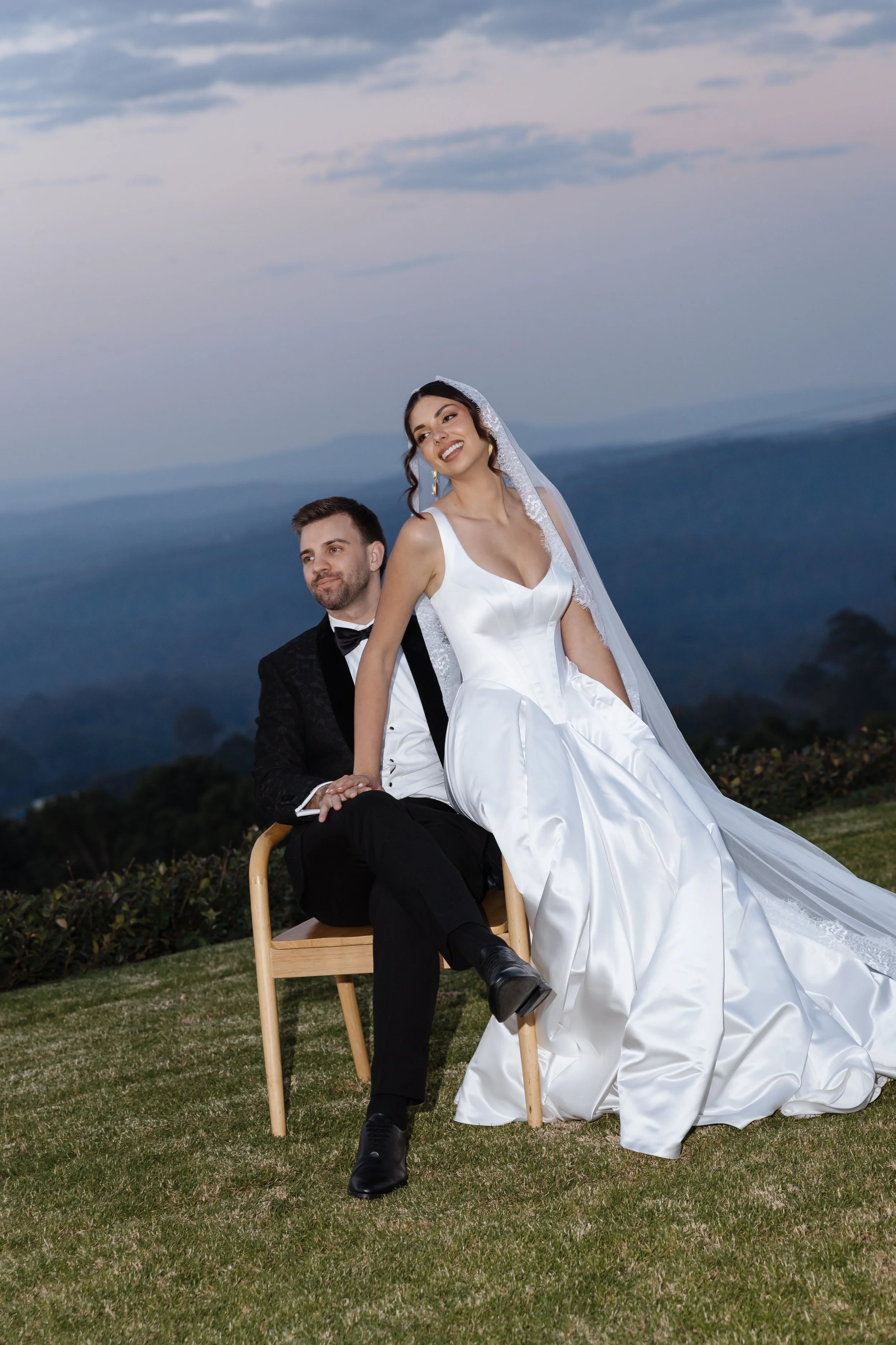 A bride and groom sitting outdoors on a chair at dusk, with the bride in a white wedding dress and veil, and the groom in a black tuxedo, with mountains and cloudy sky in the background.