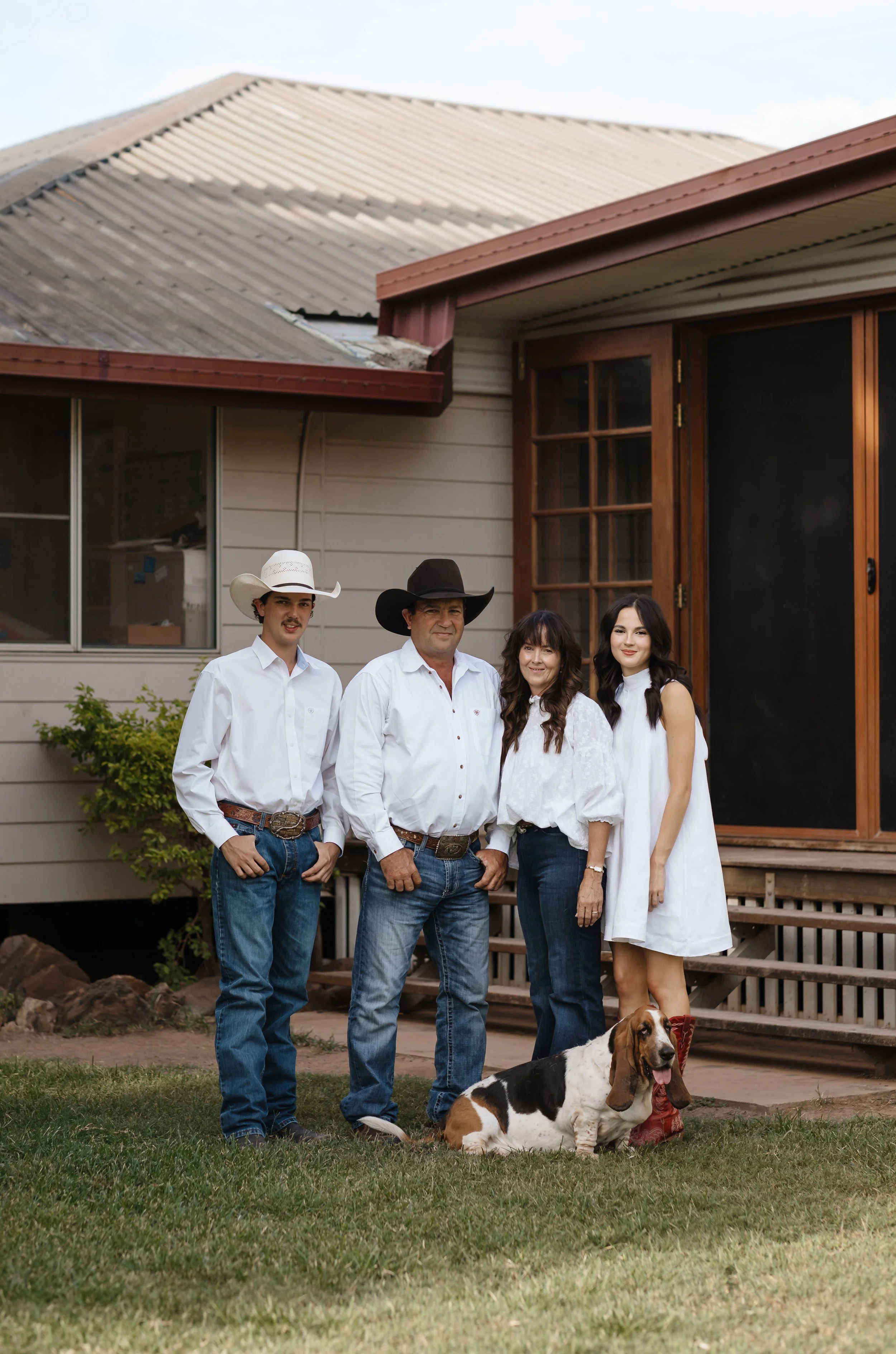 Family of four people with a dog standing in their yard in front of a house, all wearing Western or cowboy attire, including hats and jeans.