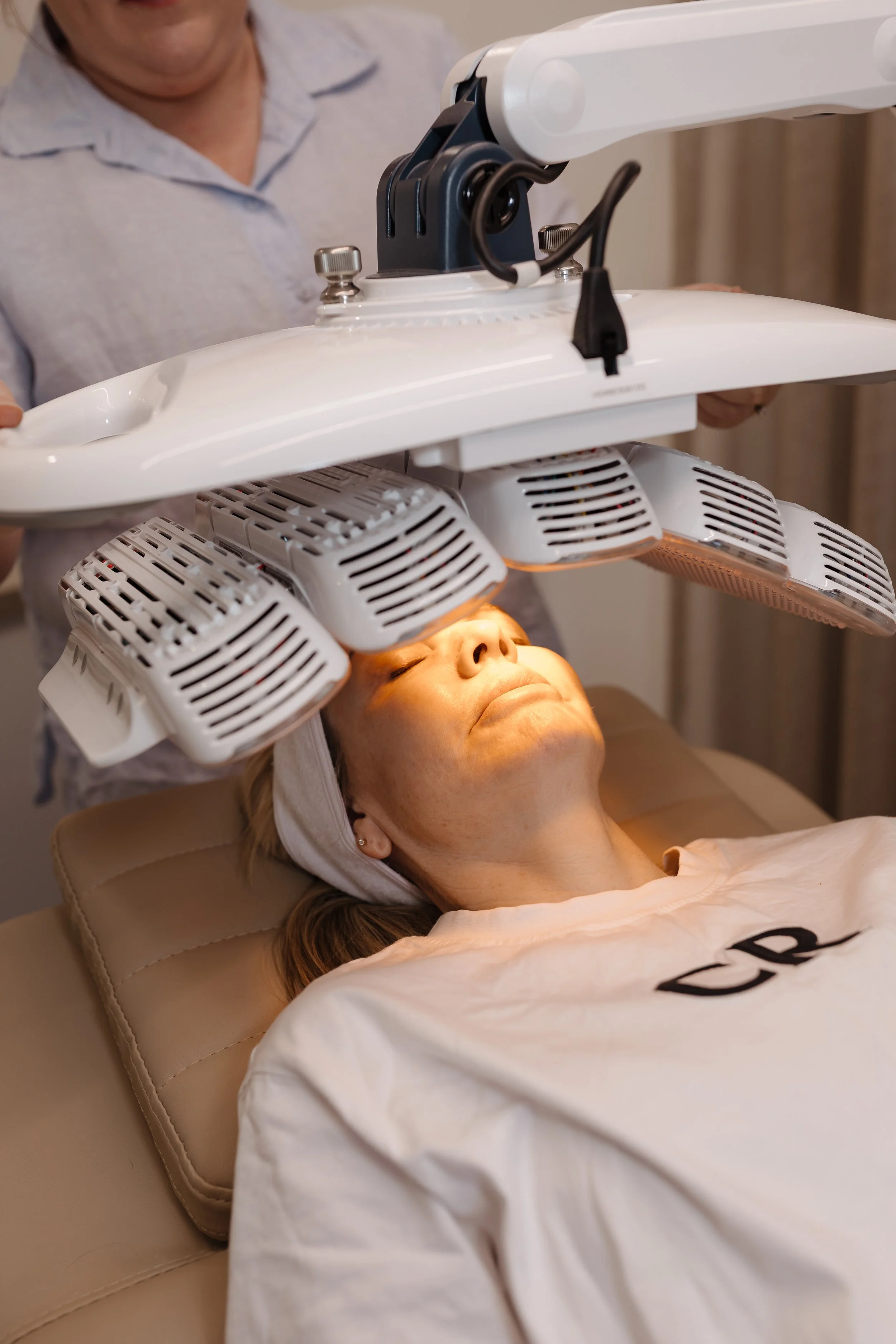 A woman lying on a treatment bed with a medical device over her face, used for a facial or skincare therapy. The device has multiple components with vents emitting warm light or heat and is positioned above her face in a clinical setting.