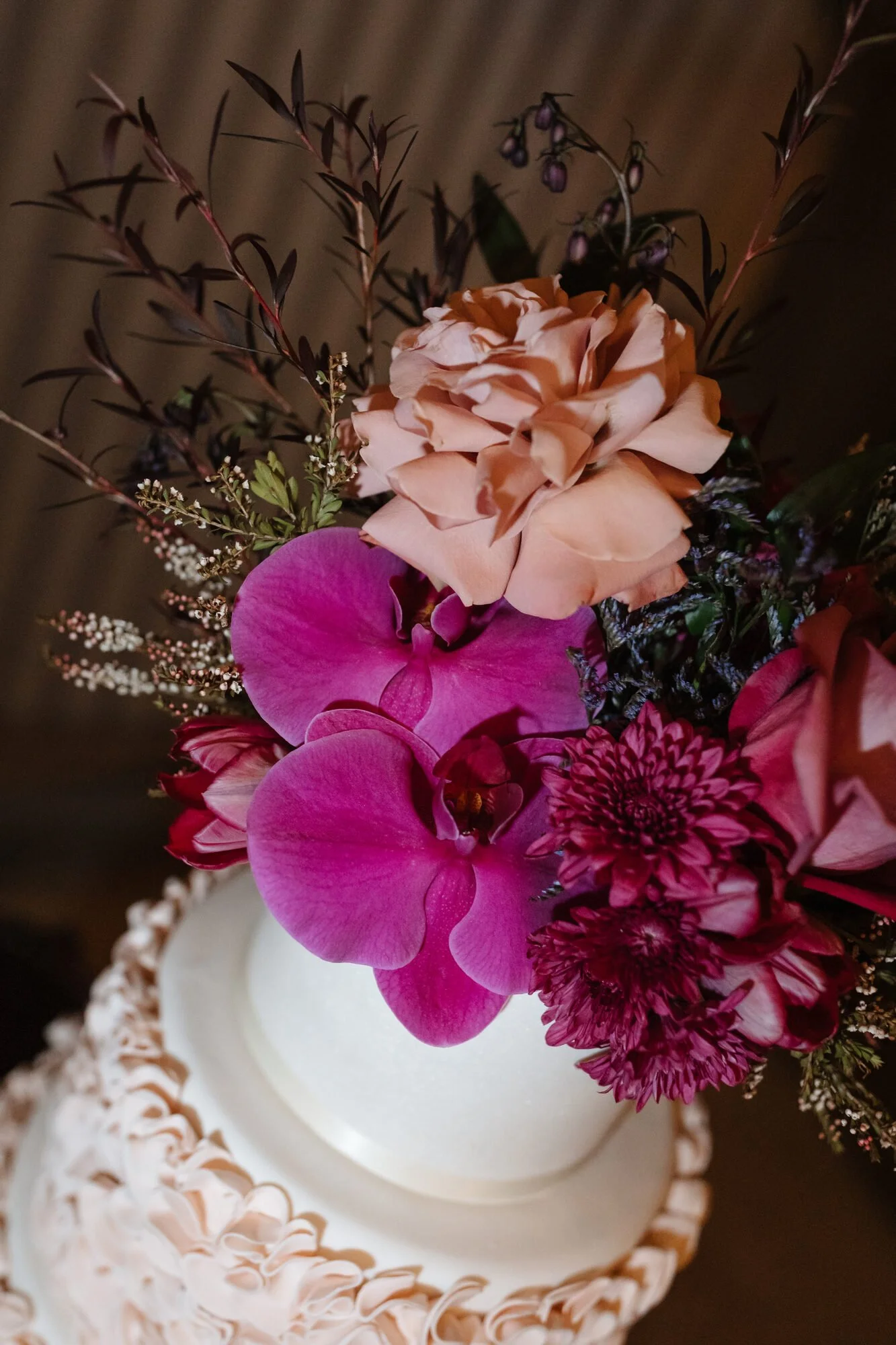 Close-up of a floral arrangement with pink, peach, purple, and white flowers in a white vase, placed on a decorative plate with a ruffled edge.