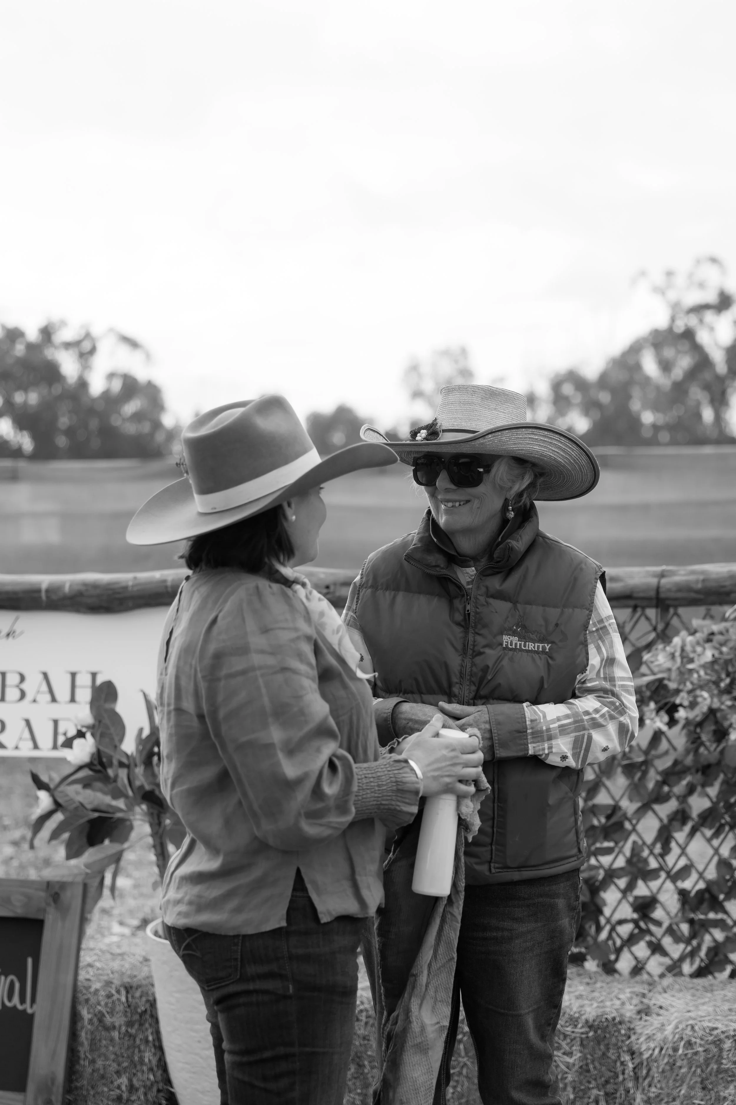Two women wearing wide-brimmed hats and sunglasses talking outdoors, with trees in the background and a sign partially visible.
