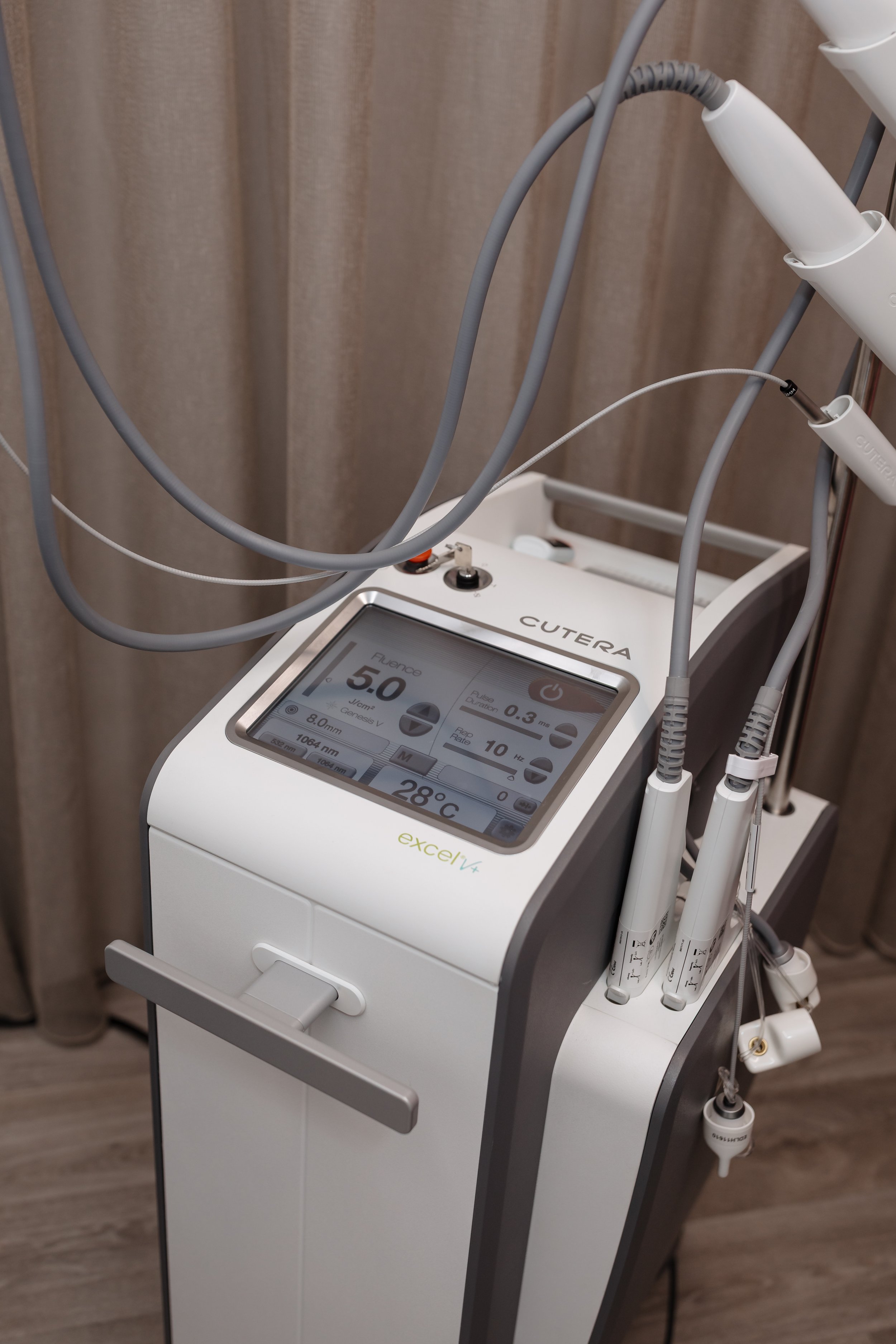 A medical or aesthetic device with a touchscreen display showing settings, connected to various handpieces and cables, positioned against a curtain background.