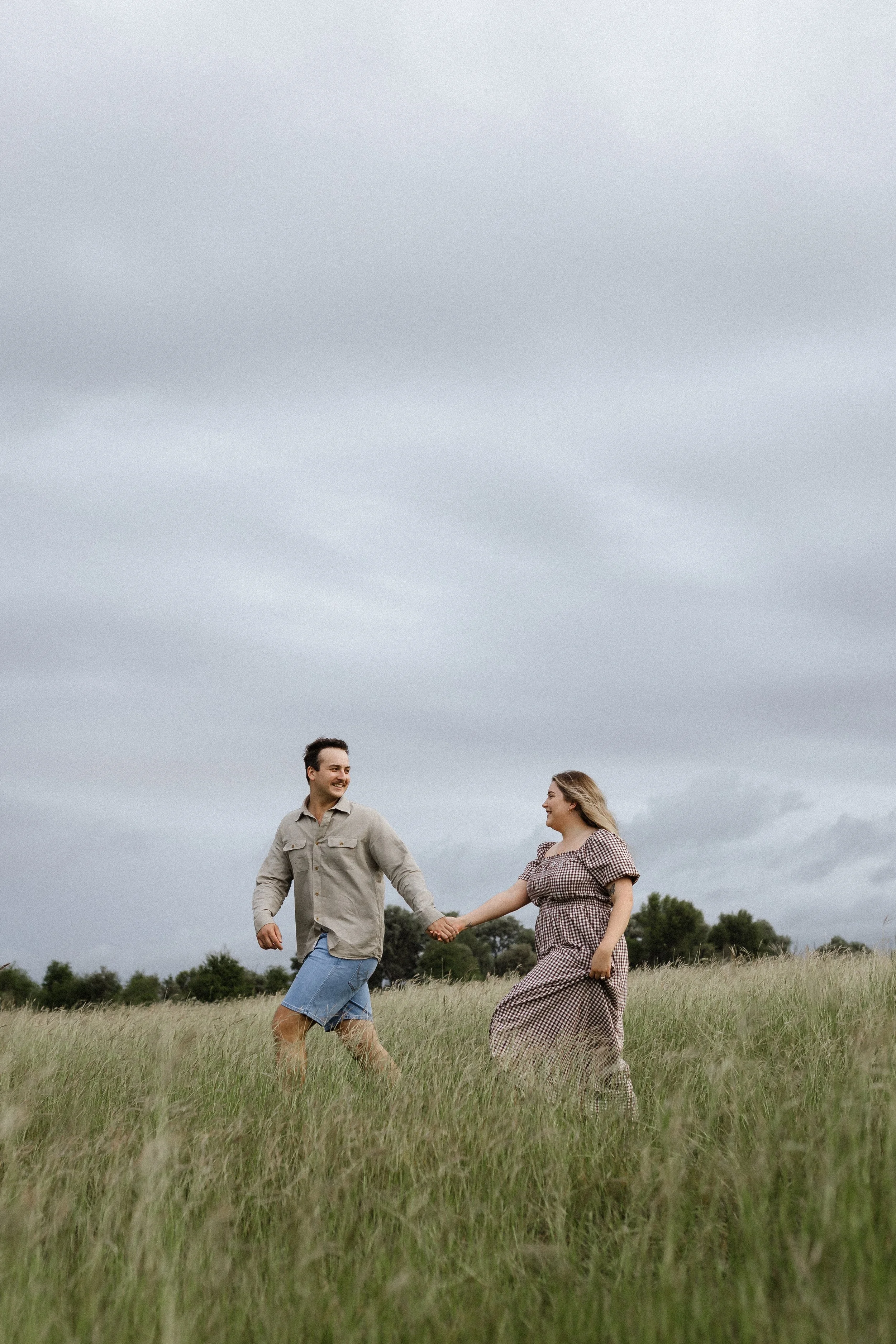 A man and woman holding hands and walking through tall grass in a field under a cloudy sky.