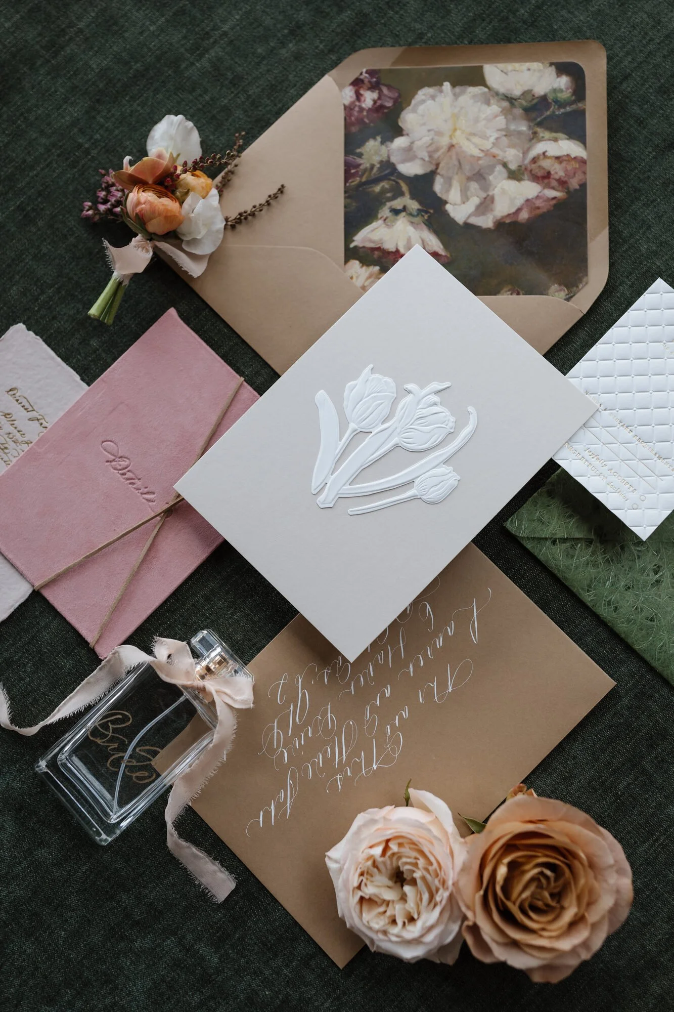 Set of wedding or greeting cards with floral designs and calligraphy, pink and cream-colored flowers, and a perfume bottle, on a dark fabric surface.