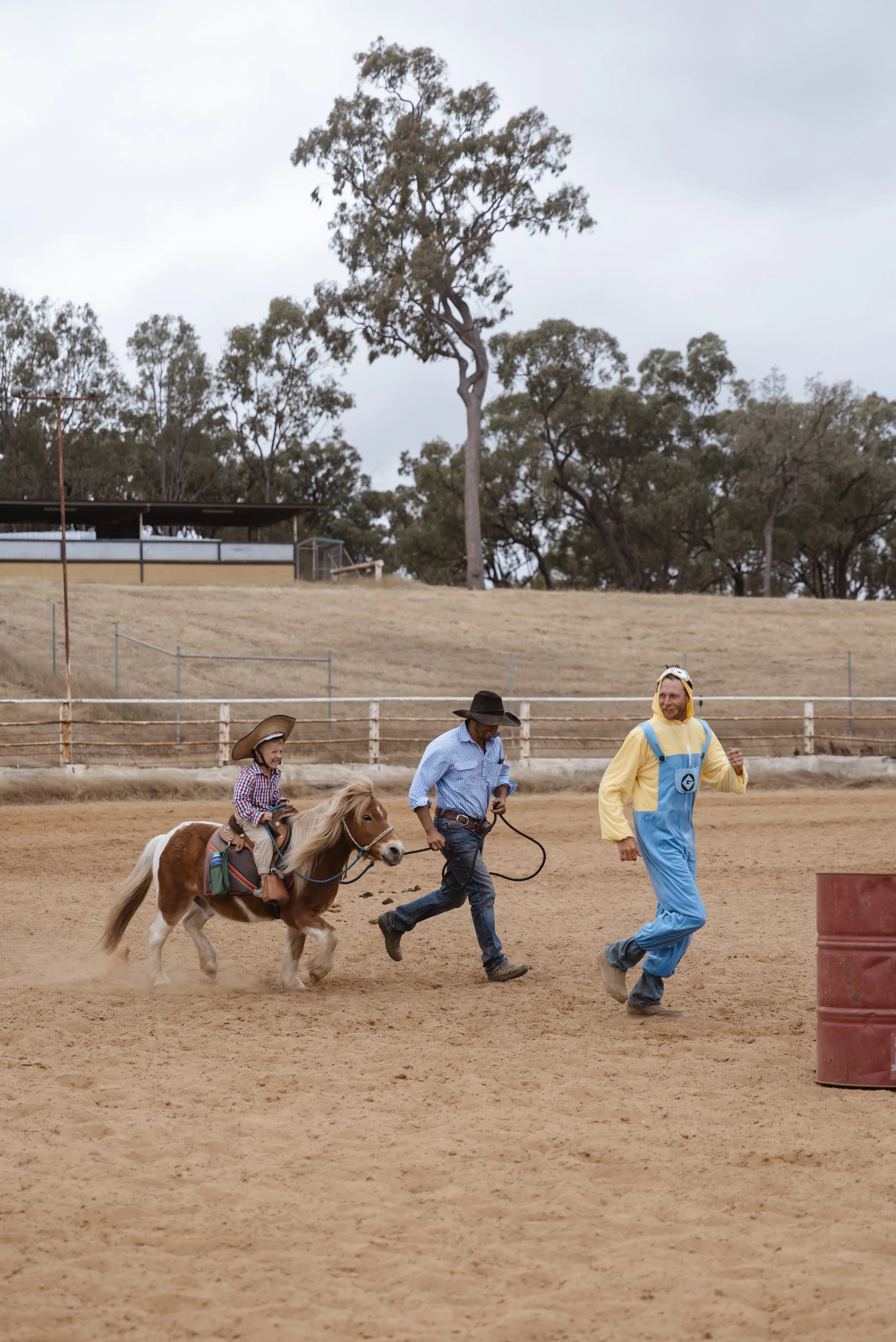 Children riding a small horse while two men walk alongside on a dirt track at an outdoor event, with trees and a building in the background.