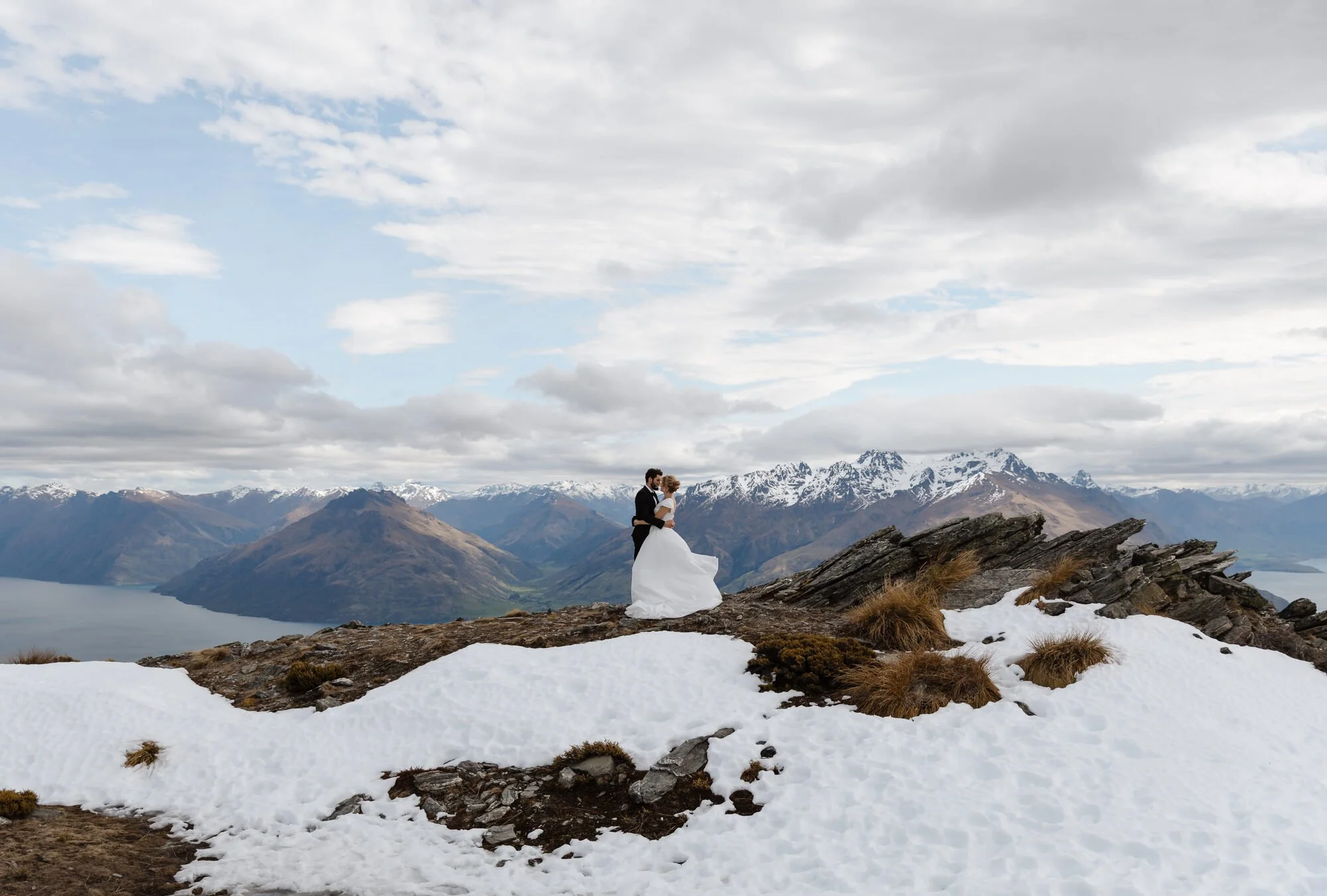 A bride and groom standing on a rocky mountaintop with snow patches, overlooking a valley and snow-capped mountains under a cloudy sky.