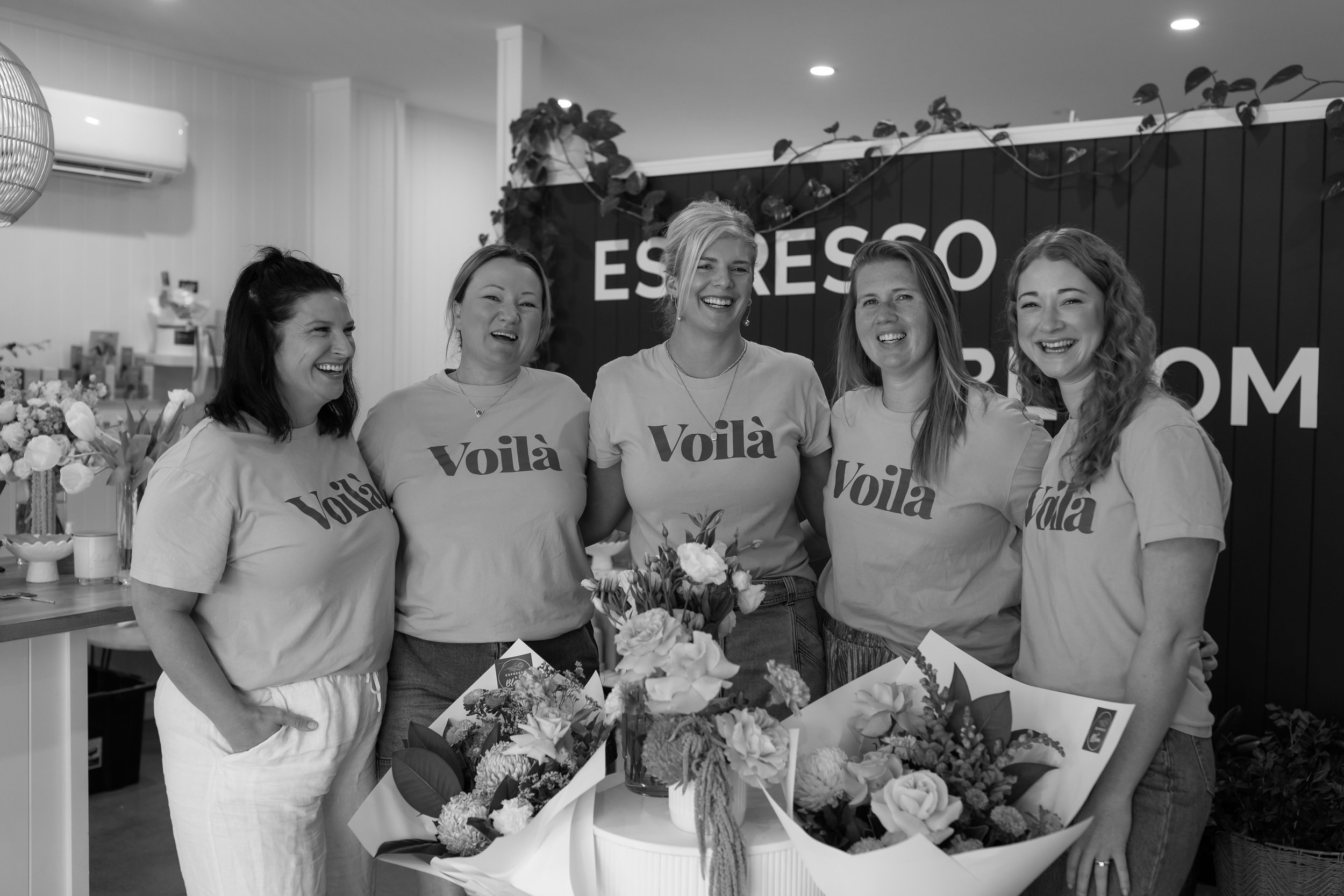 Five women standing behind a table of flowers, smiling, in a room with a sign that reads 'Espresso'. All are wearing T-shirts with the word 'Voilà' printed on them.