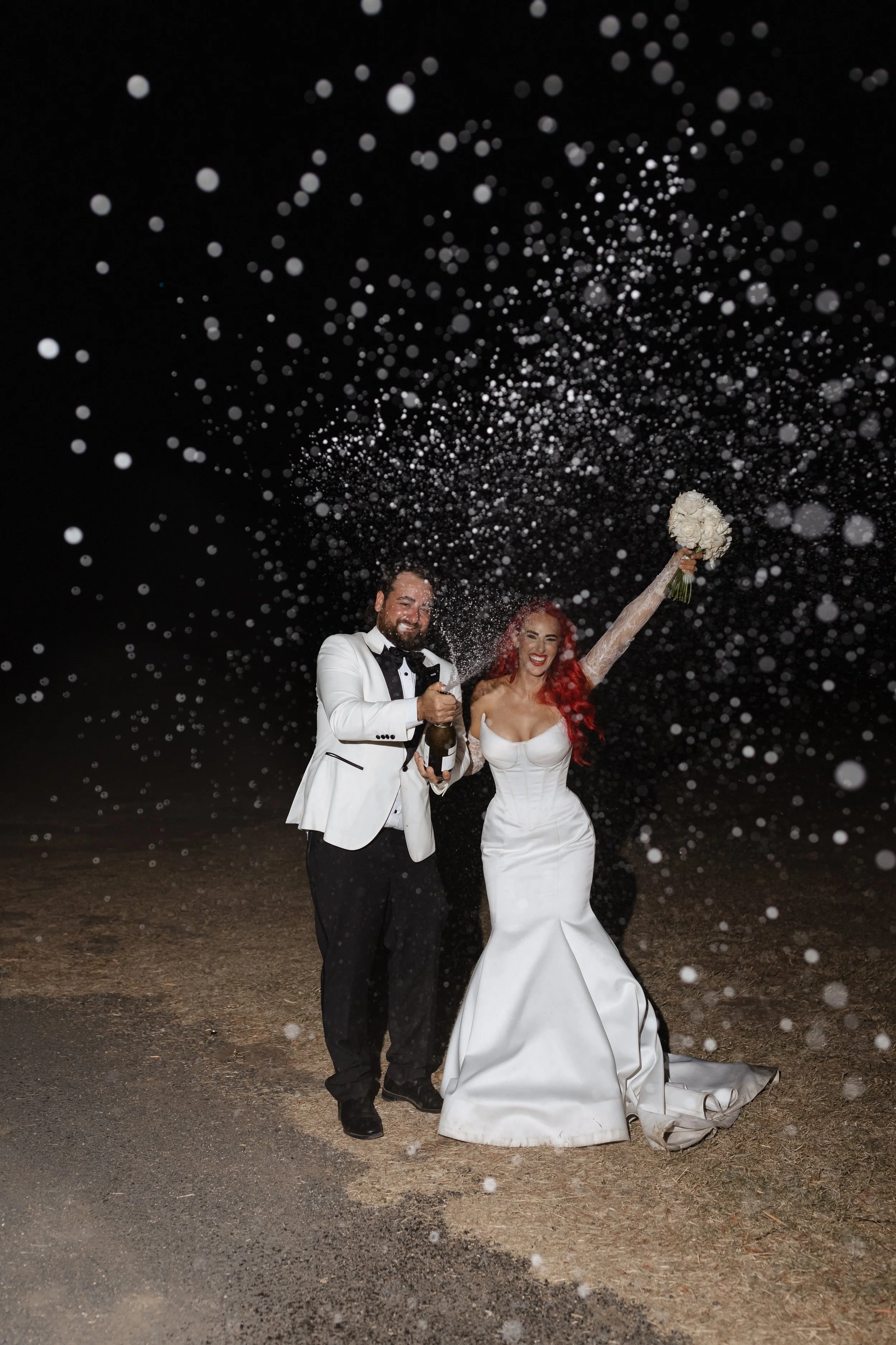 A newly married couple celebrating outdoors at night, with white confetti falling around them. The bride, wearing a strapless white wedding dress, holds a bouquet of white flowers in one hand and raises her other arm joyfully. The groom, dressed in a