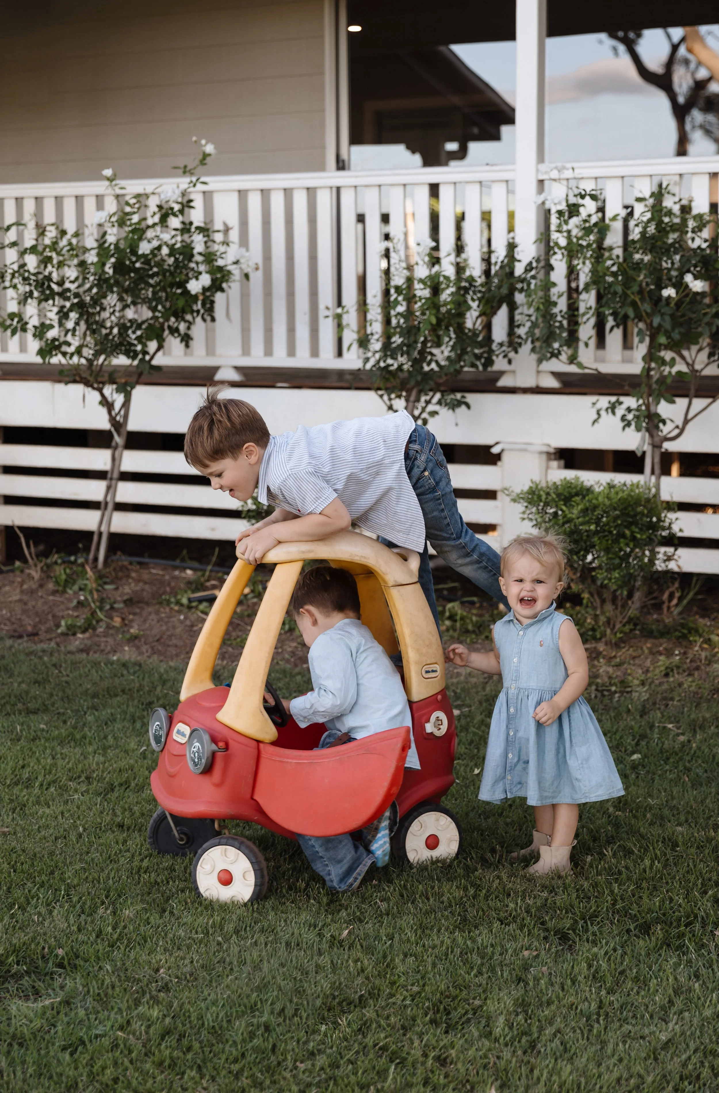 Children playing in a backyard with a play vehicle. A boy in a striped shirt climbs on top of the play vehicle, while a girl in a denim dress stands beside it, crying. Two other children are inside the vehicle. The background features a white fence, 