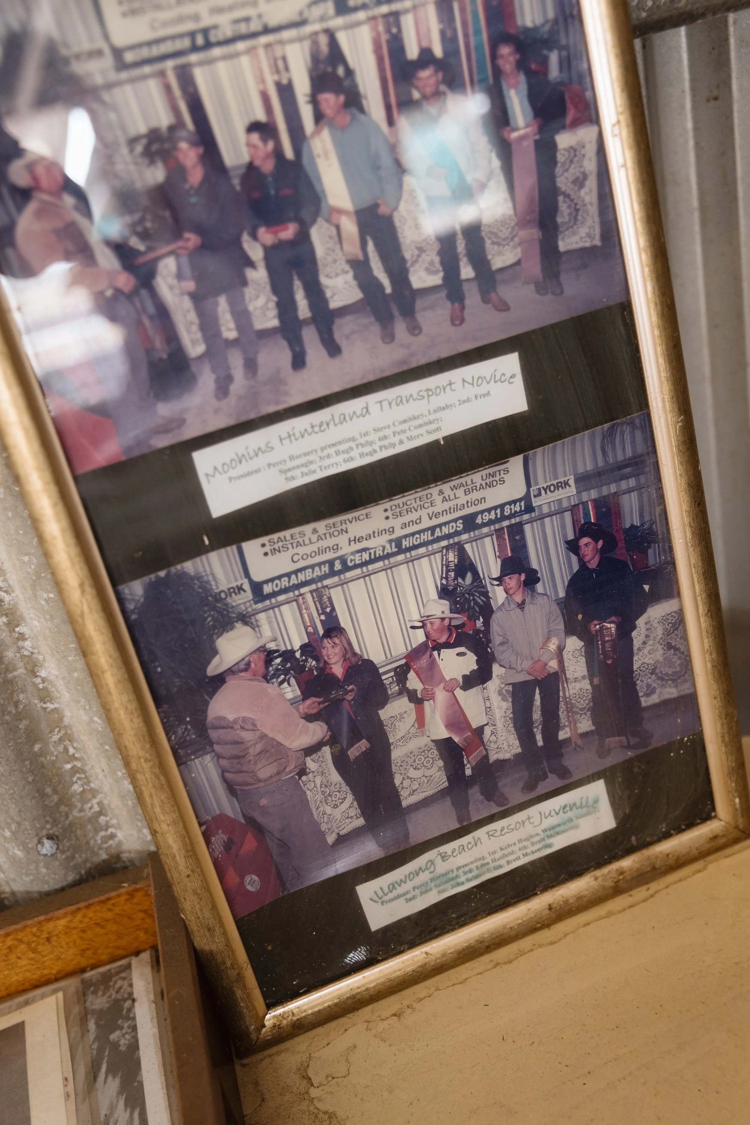 Group of people standing in a row, some with cowboy hats, in front of a decorated wall with striped and patterned fabric and banners, during an event titled 'Moohins Hinterland Transport Novice'.