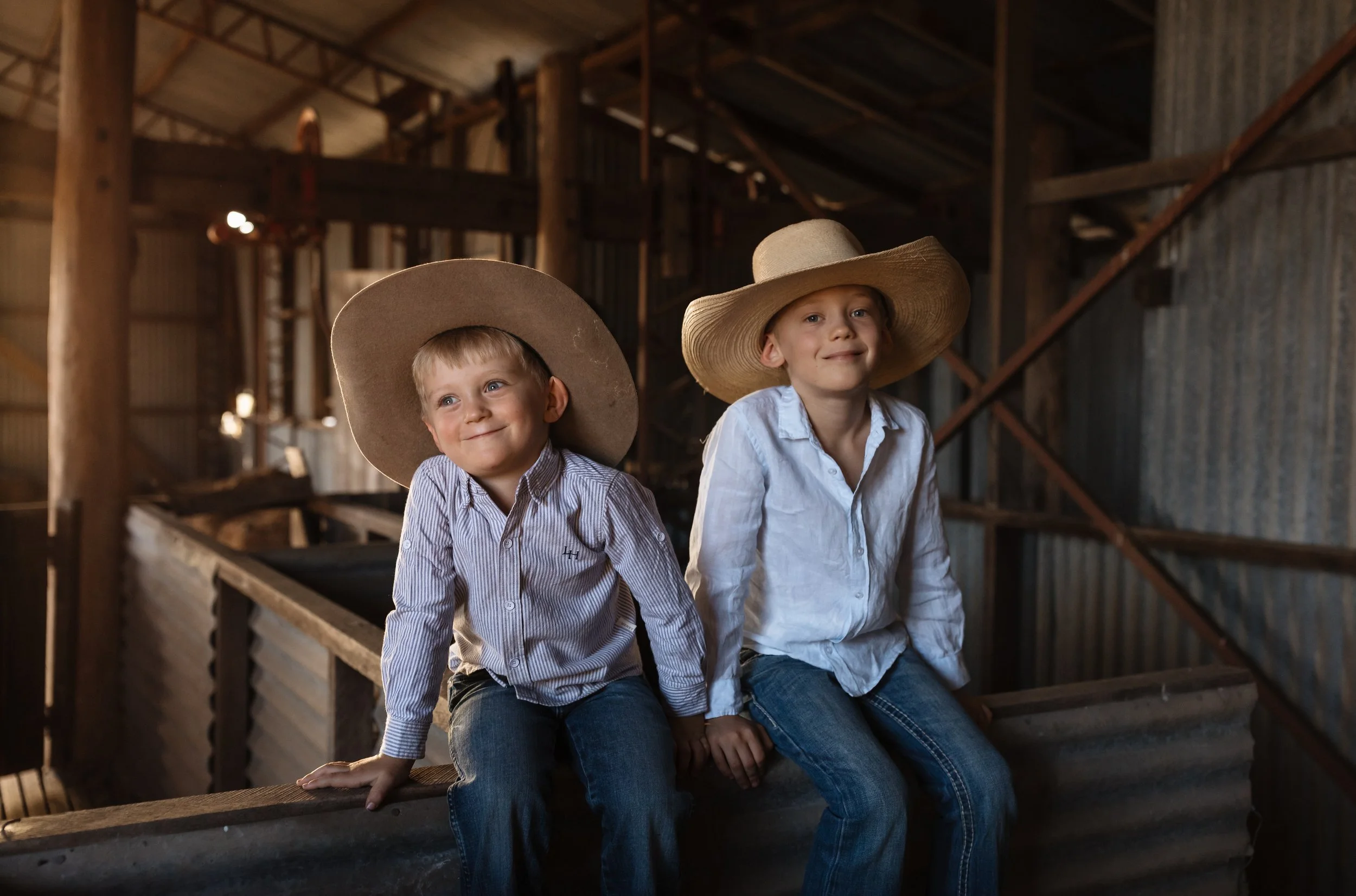 Two young boys sitting on a wooden barn ledge, wearing large cowboy hats and casual shirts, smiling at the camera.