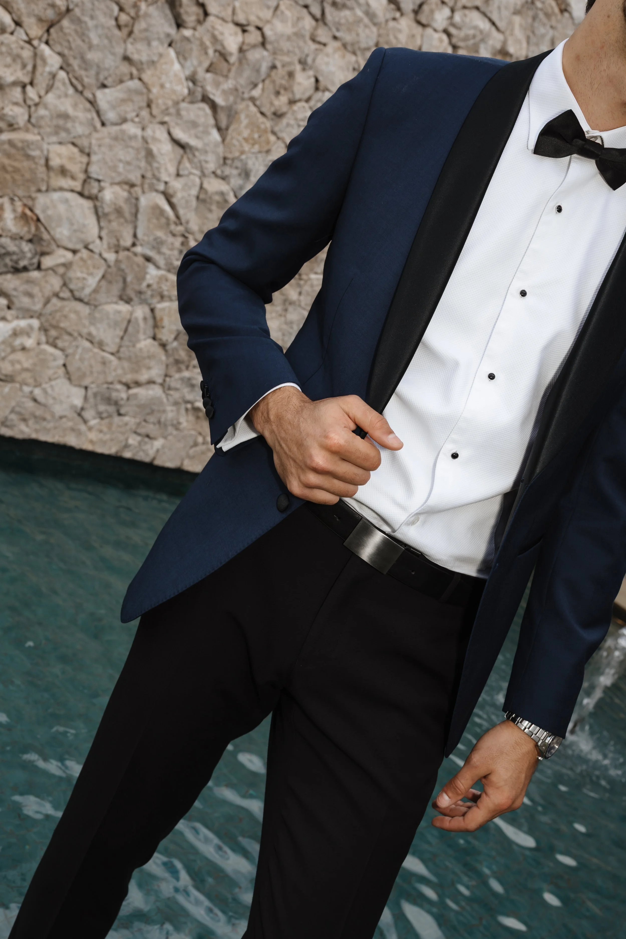 Man dressed in a tuxedo with a black bow tie, white shirt, and blue jacket, standing near water with a stone wall in the background.