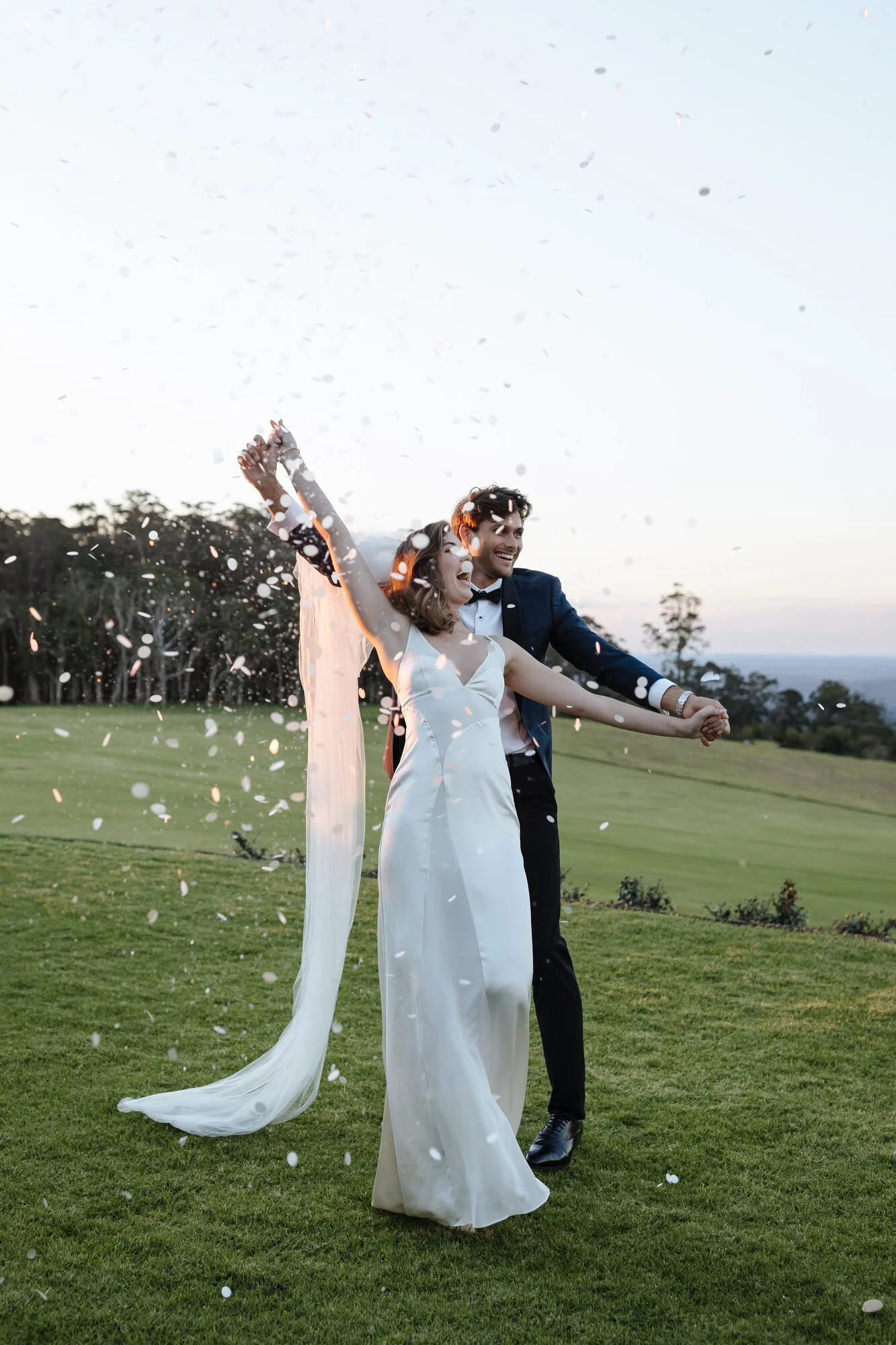A happy couple in wedding attire celebrating outdoors on a grassy field with trees and a distant view of the ocean or lake in the background. The woman is wearing a white wedding dress with a long train, and the man is in a navy blue suit. They are h