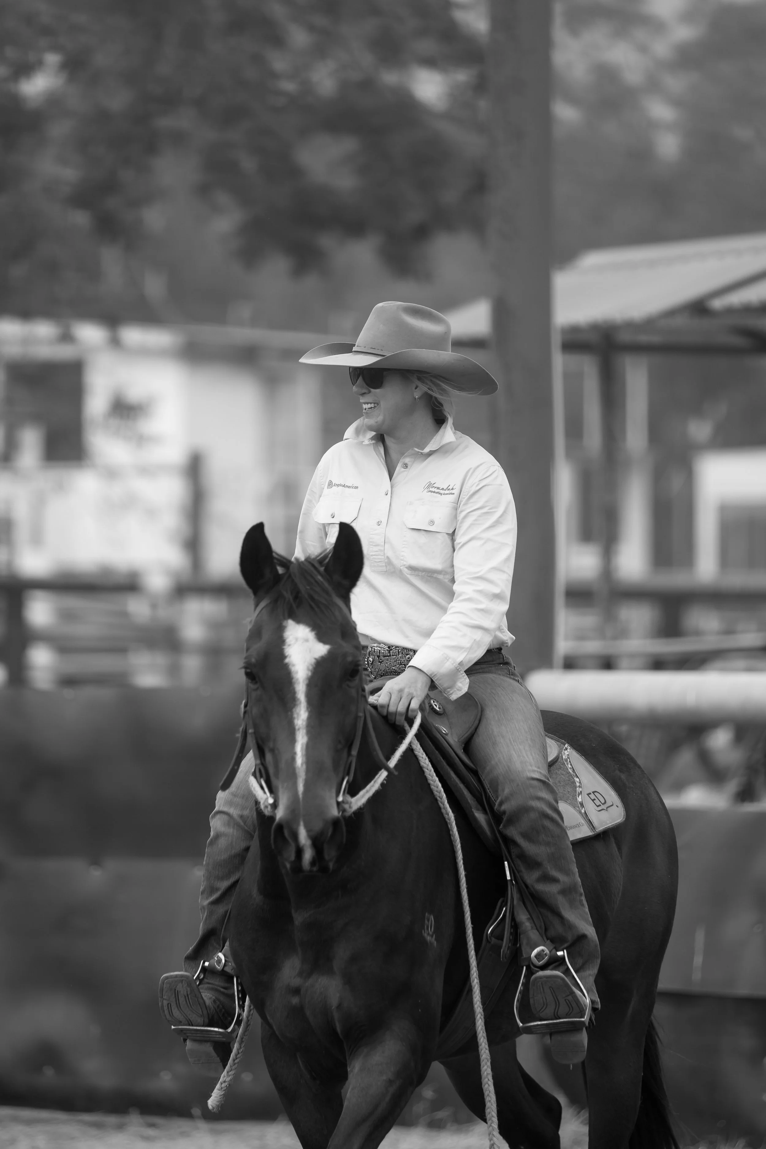 A woman riding a horse outdoors, wearing a wide-brimmed hat, sunglasses, and a button-up shirt, smiling as she rides.