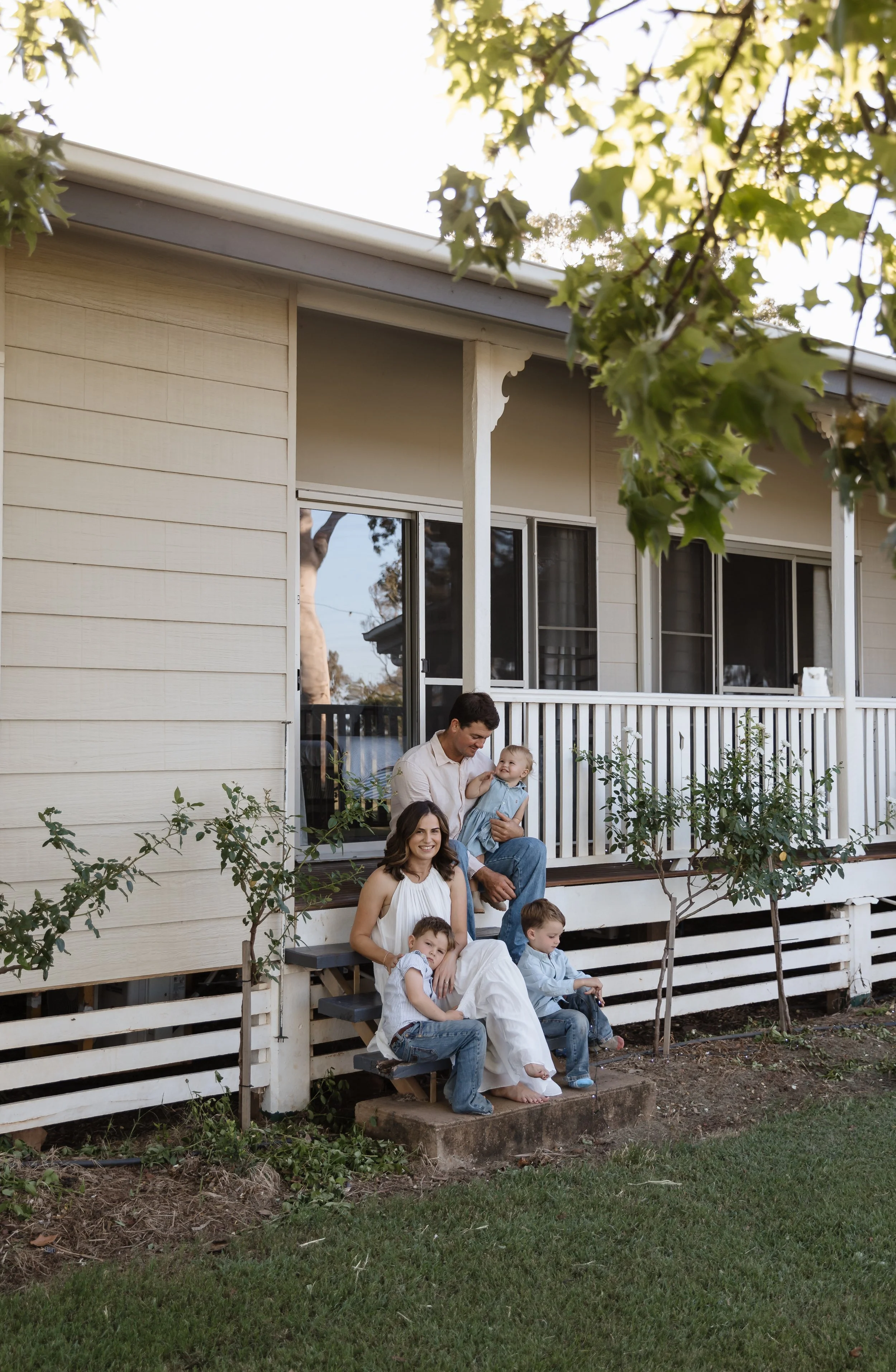 A family of five, including two adult women and a man with three young children, sitting on steps outside a house with light beige siding, a small porch, and trees and greenery around.