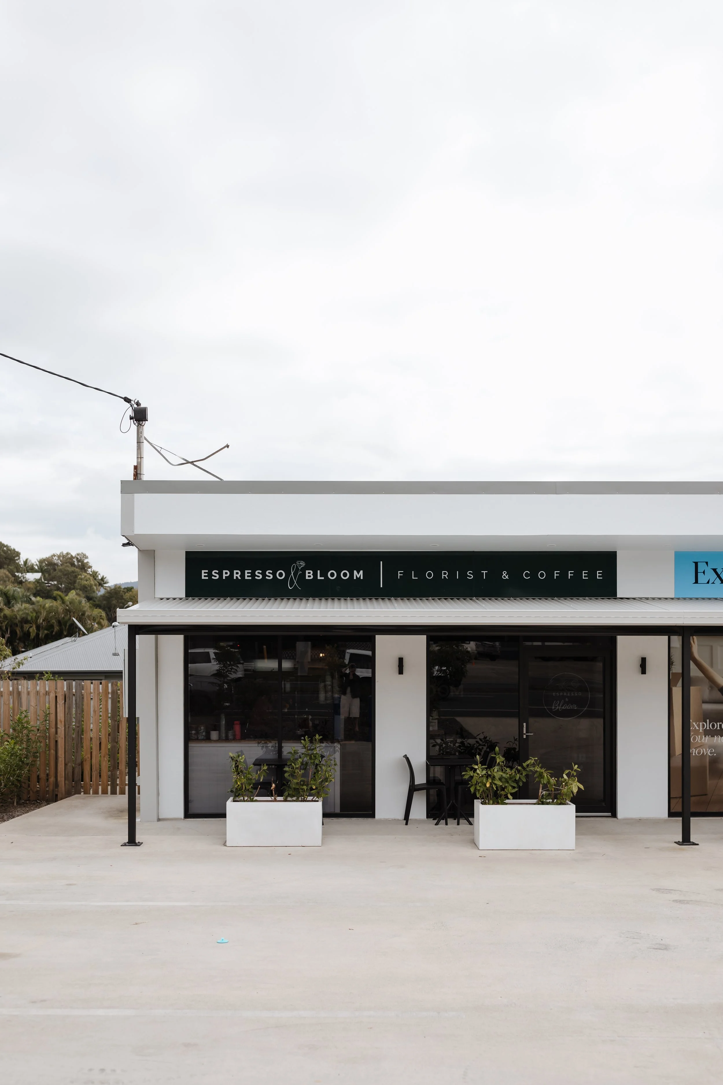 Front view of a modern coffee shop with outdoor seating, black sign reading 'Espresso & Bloom Florist & Coffee', and a gray overcast sky.
