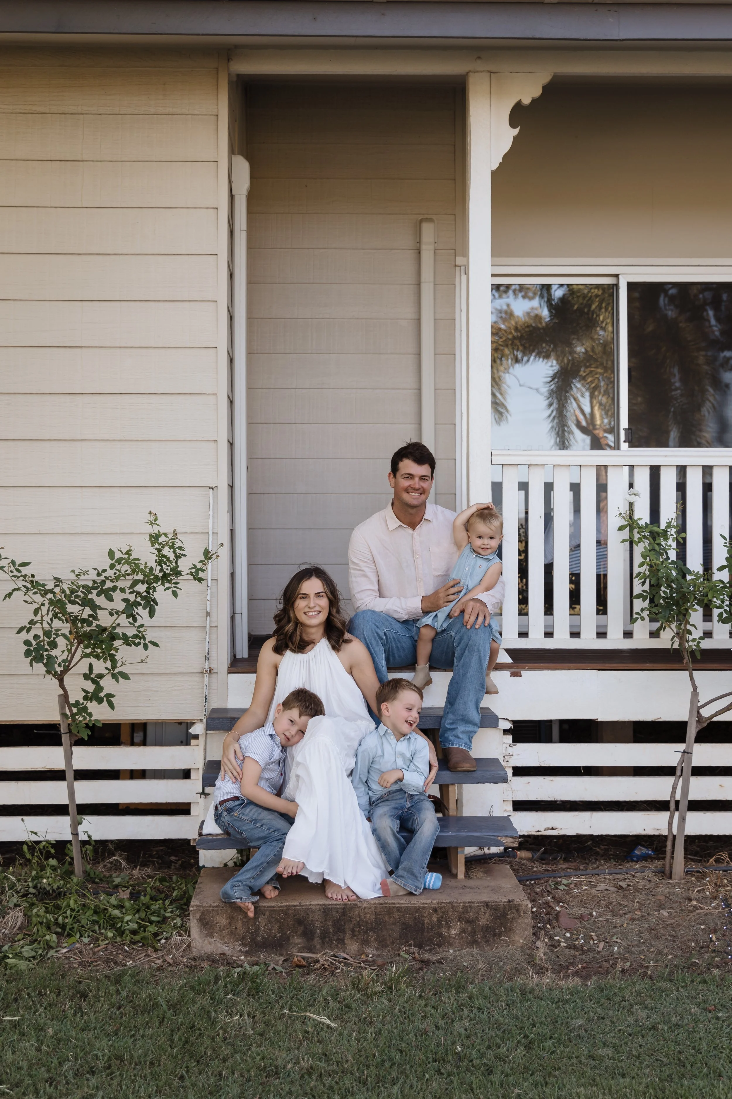 A family of six posing on the steps of a porch in front of a beige house with white trim and a large window, with trees and blue sky in the background.