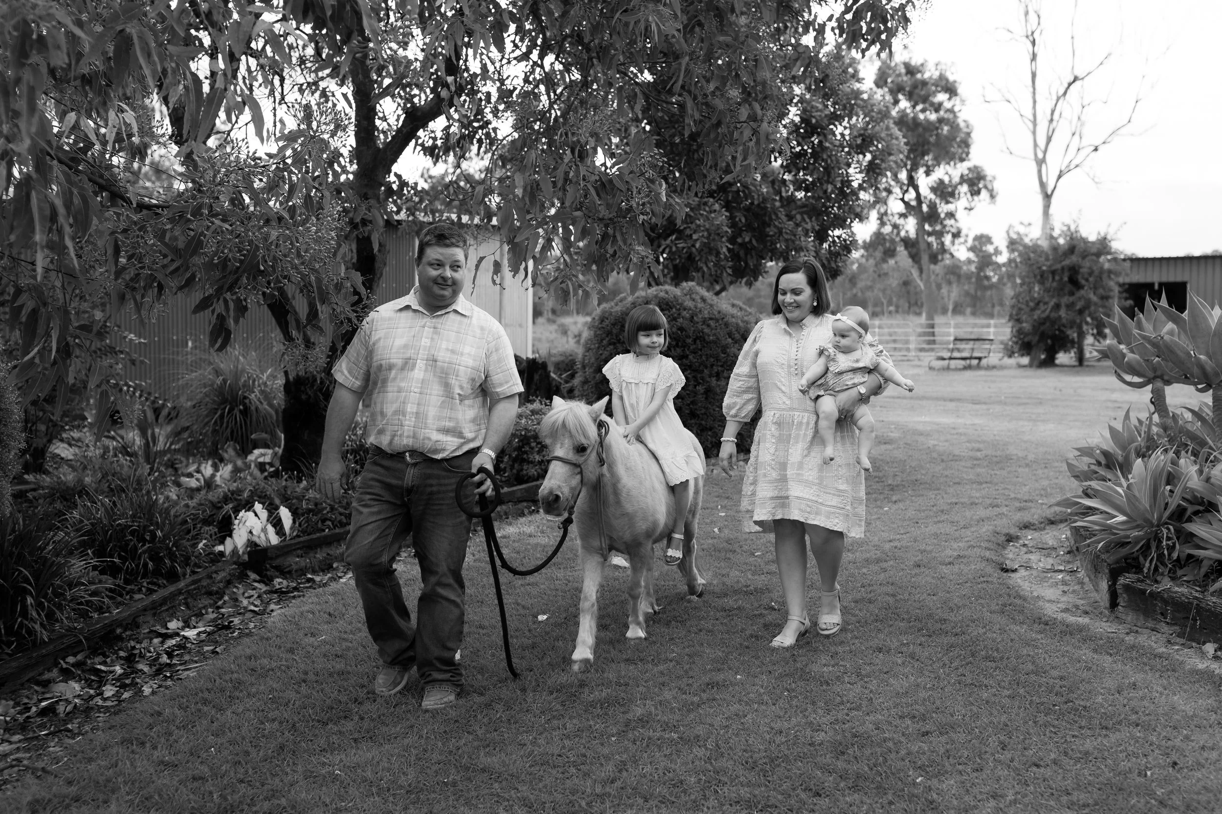 A black and white photo of a family walking in a garden. A man is leading a pony, with a young girl sitting on its back, while a woman holds a baby and walks beside them.