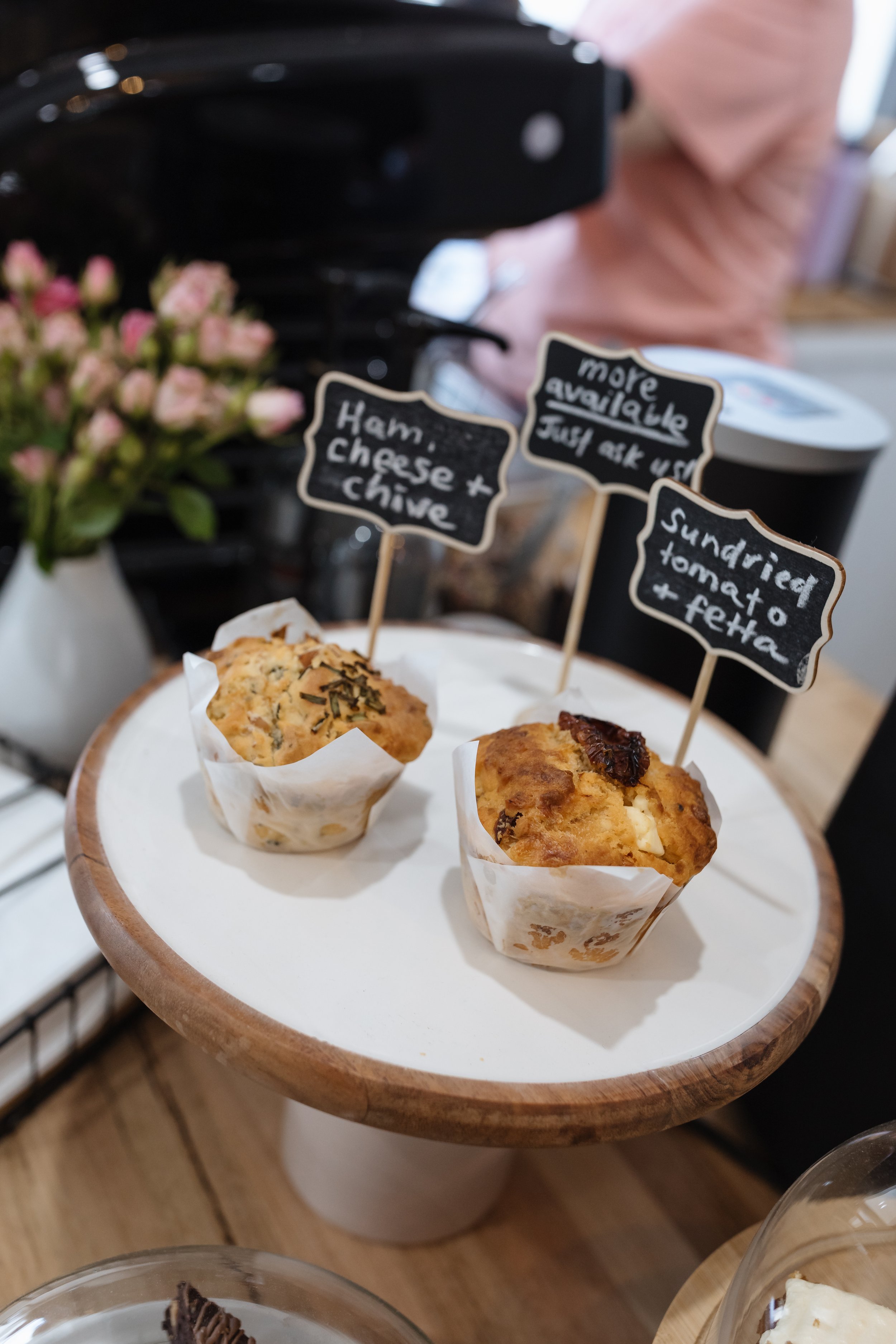 Two muffins on a white round tray, one flavored with cheese and herbs, the other with dried fruit or chocolate chips, with small chalkboard signs labeling each muffin.