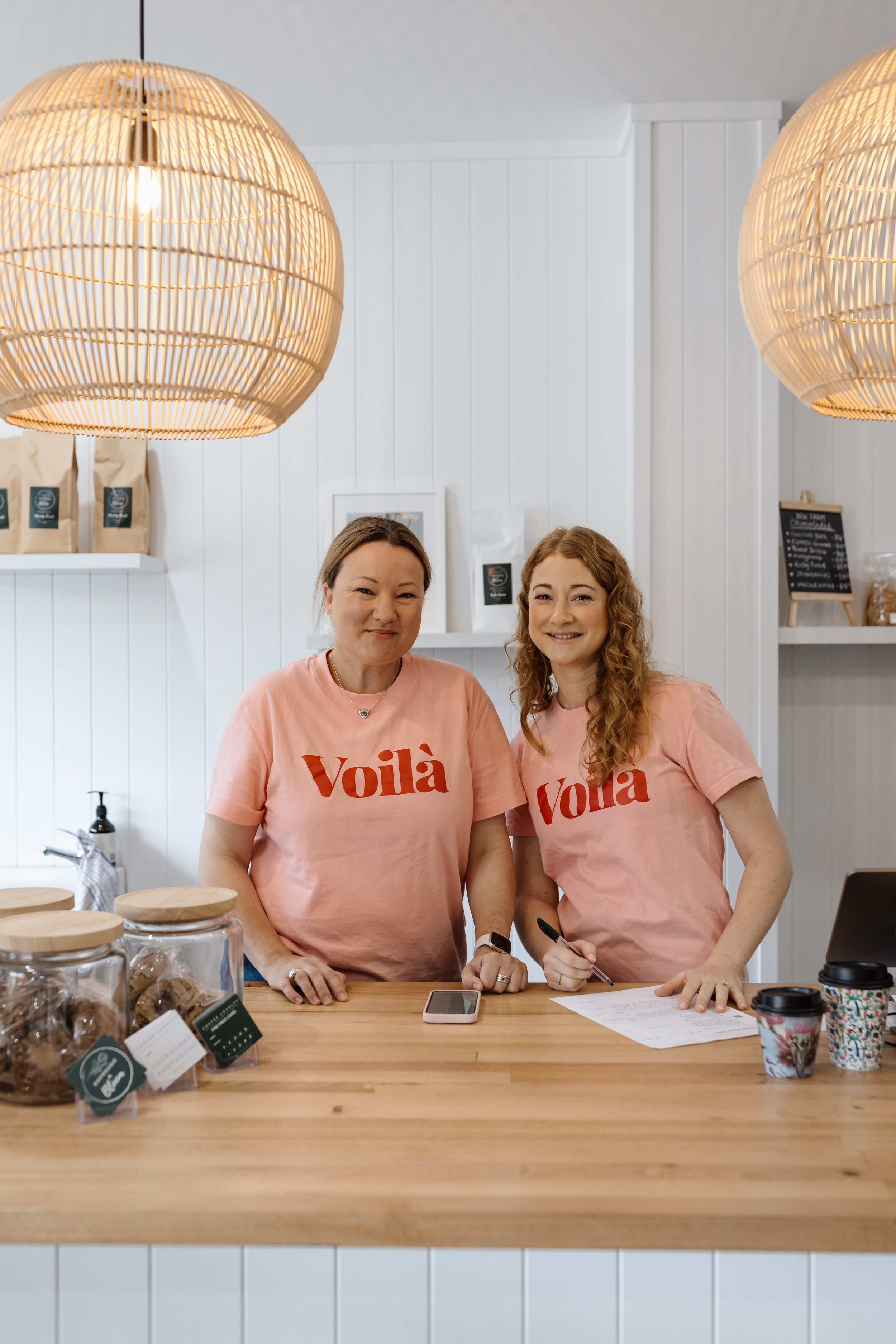 Two women standing behind a wooden counter in a cafe, both wearing pink T-shirts with the word "Voilà" printed in red, smiling at the camera. The cafe has white paneled walls, hanging pendant lights, jars of baked goods, and a menu board in the backg