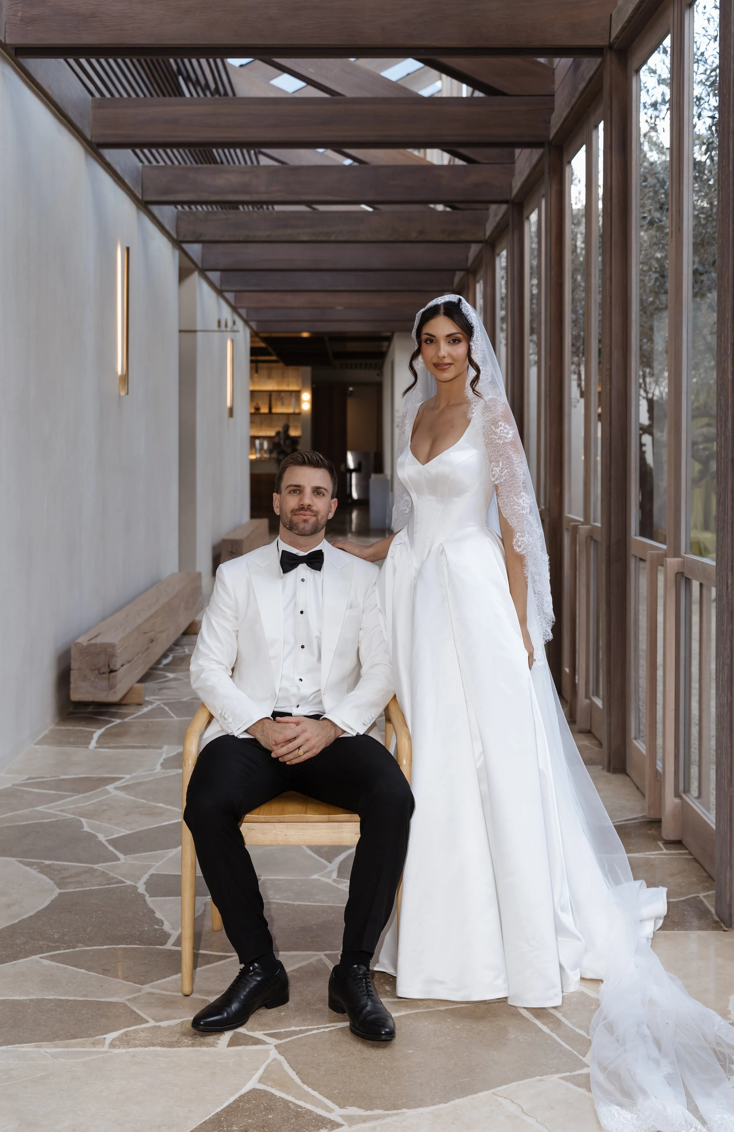 A bride and groom in wedding attire posing indoors, with the bride standing and the groom seated on a wooden chair.