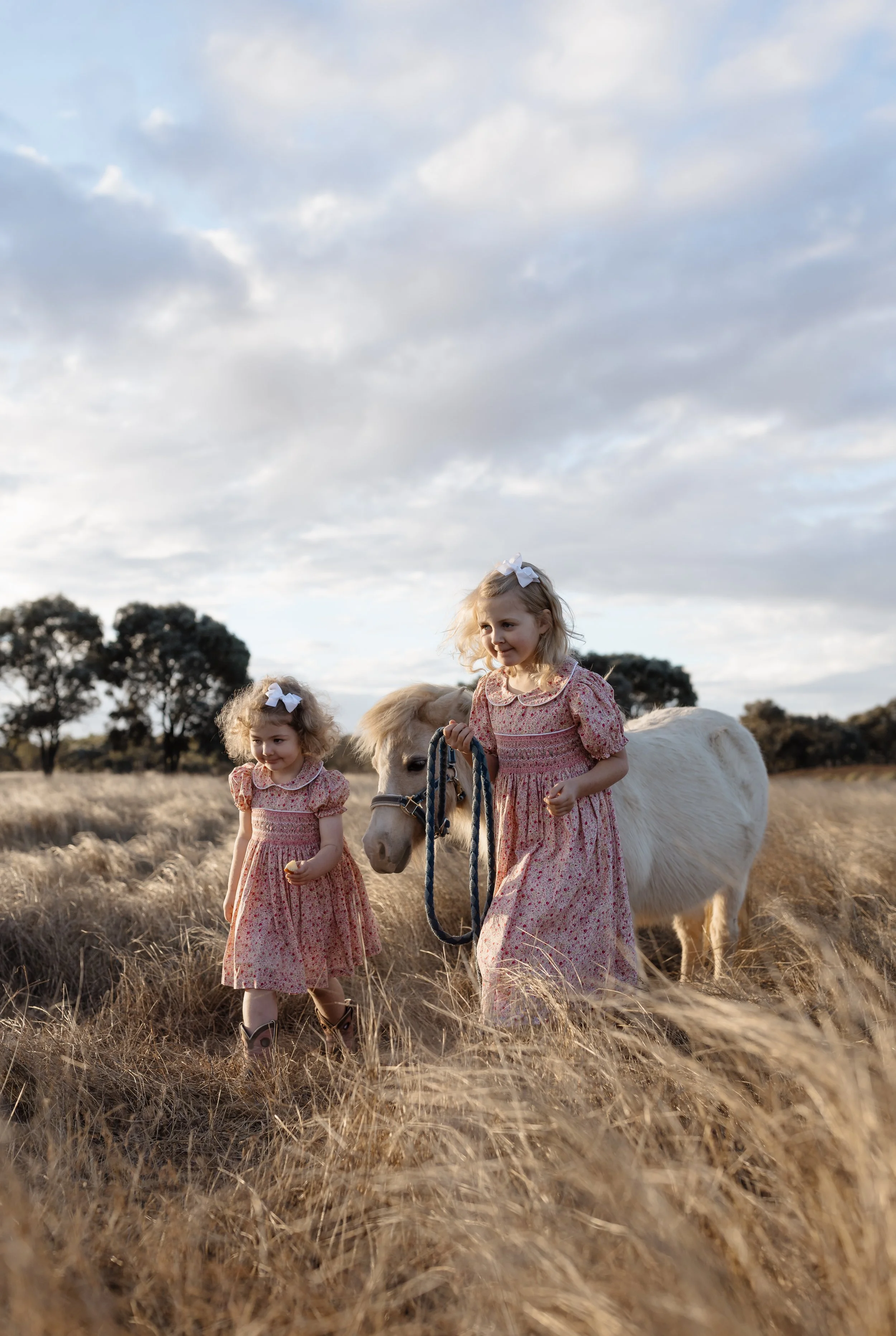 Two young girls in matching floral dresses and white hair bows, each holding a piece of grass, walking in a field with a small white pony. The girl on the right is holding the pony's halter, and a few trees are visible in the background against a clo