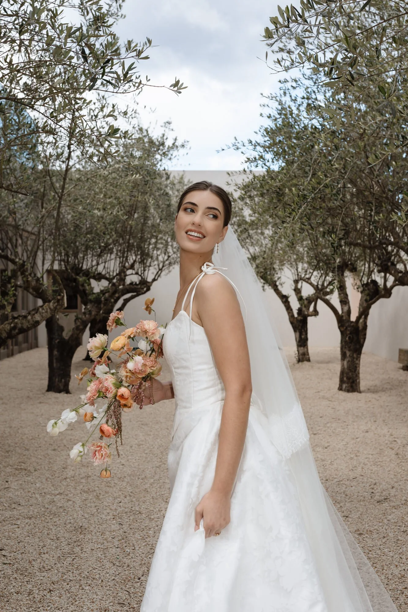 A bride in a white wedding dress holding a bouquet of flowers, standing outdoors in front of trees with green foliage on a cloudy day.