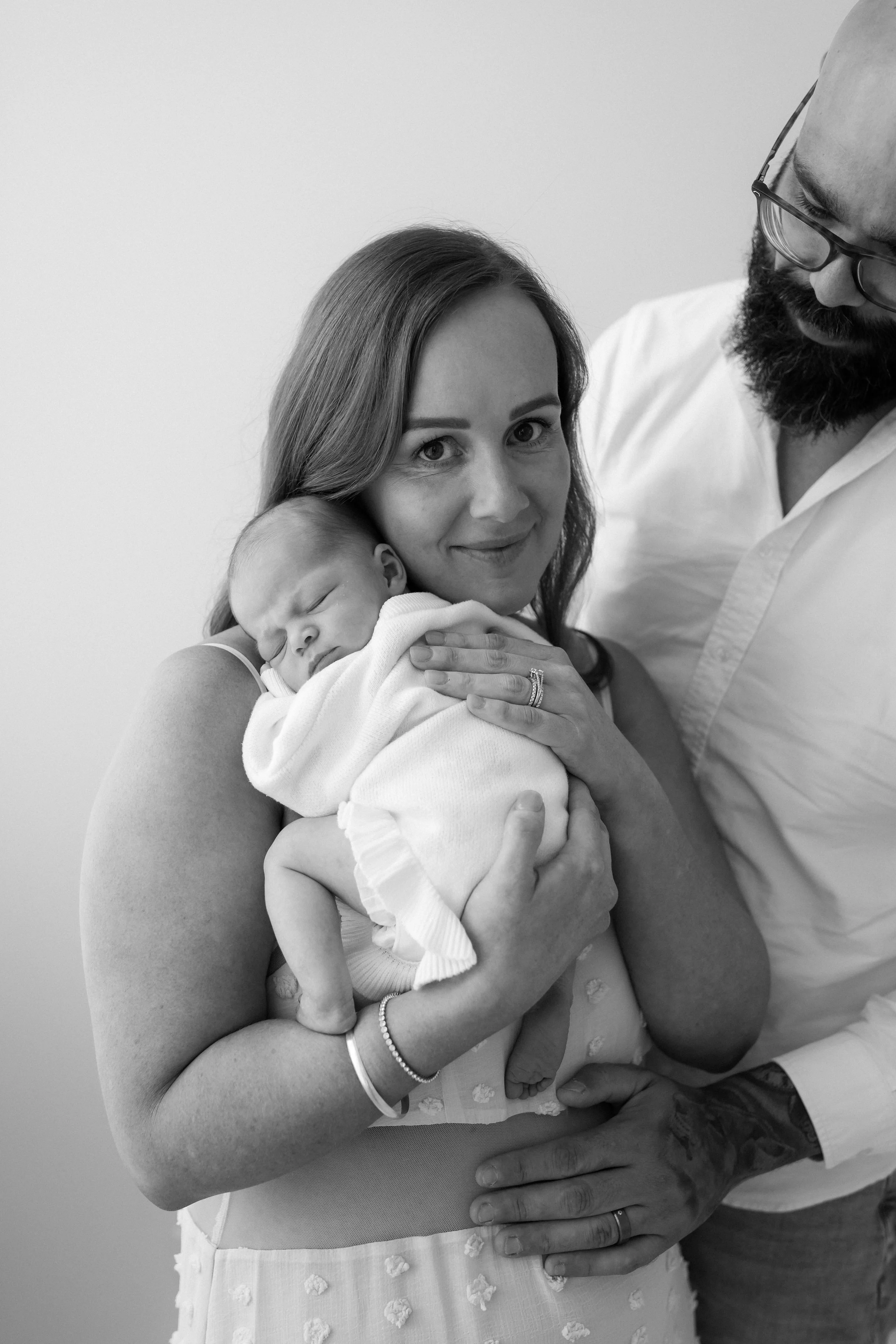 A woman holding a newborn baby, with a man standing nearby, all in black and white. The woman is smiling, the baby is sleeping.