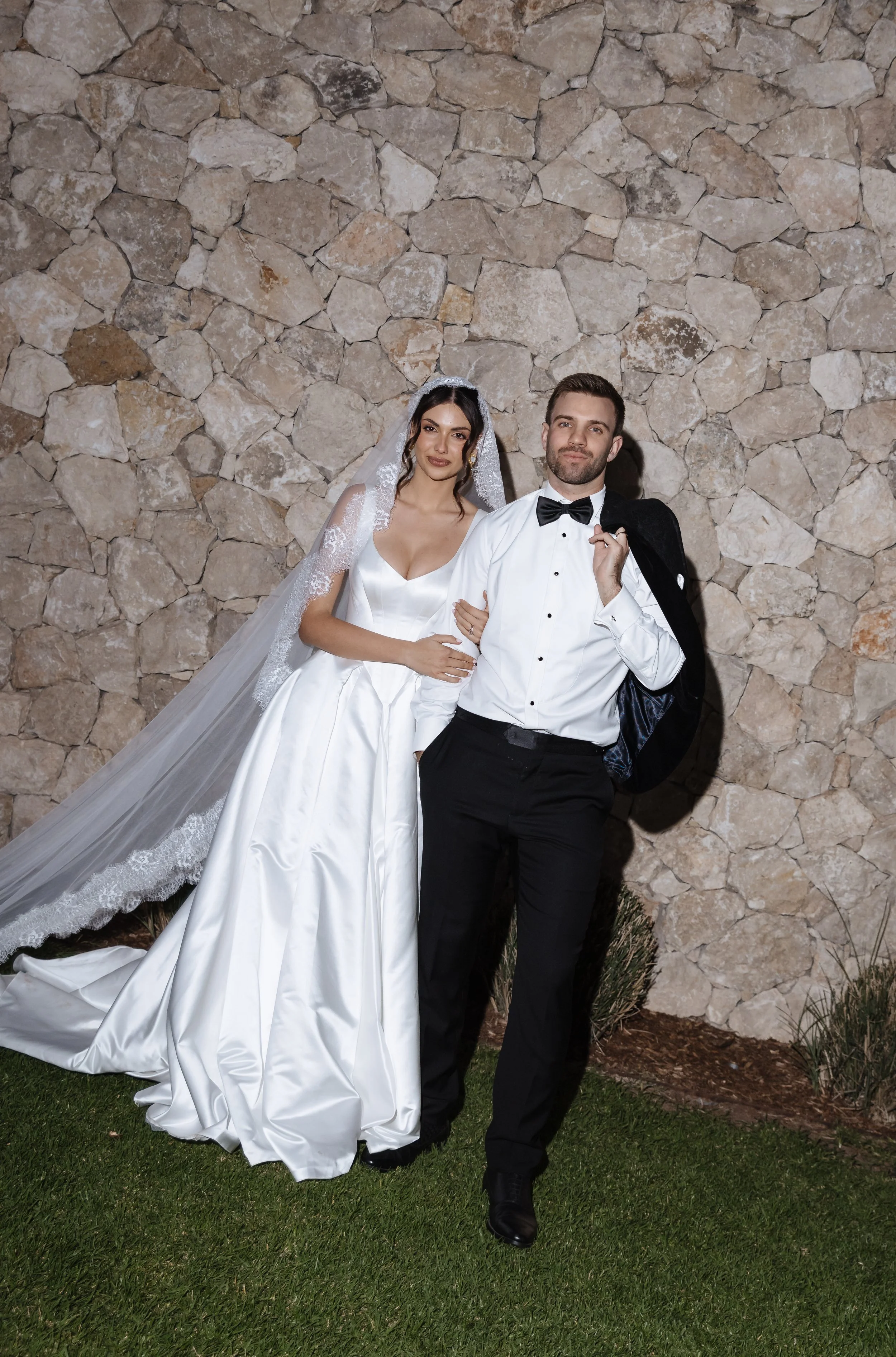 A bride and groom pose together outdoors in front of a stone wall. The bride wears a white wedding gown with a veil, and the groom wears a tuxedo with a bow tie, holding a jacket over his shoulder.
