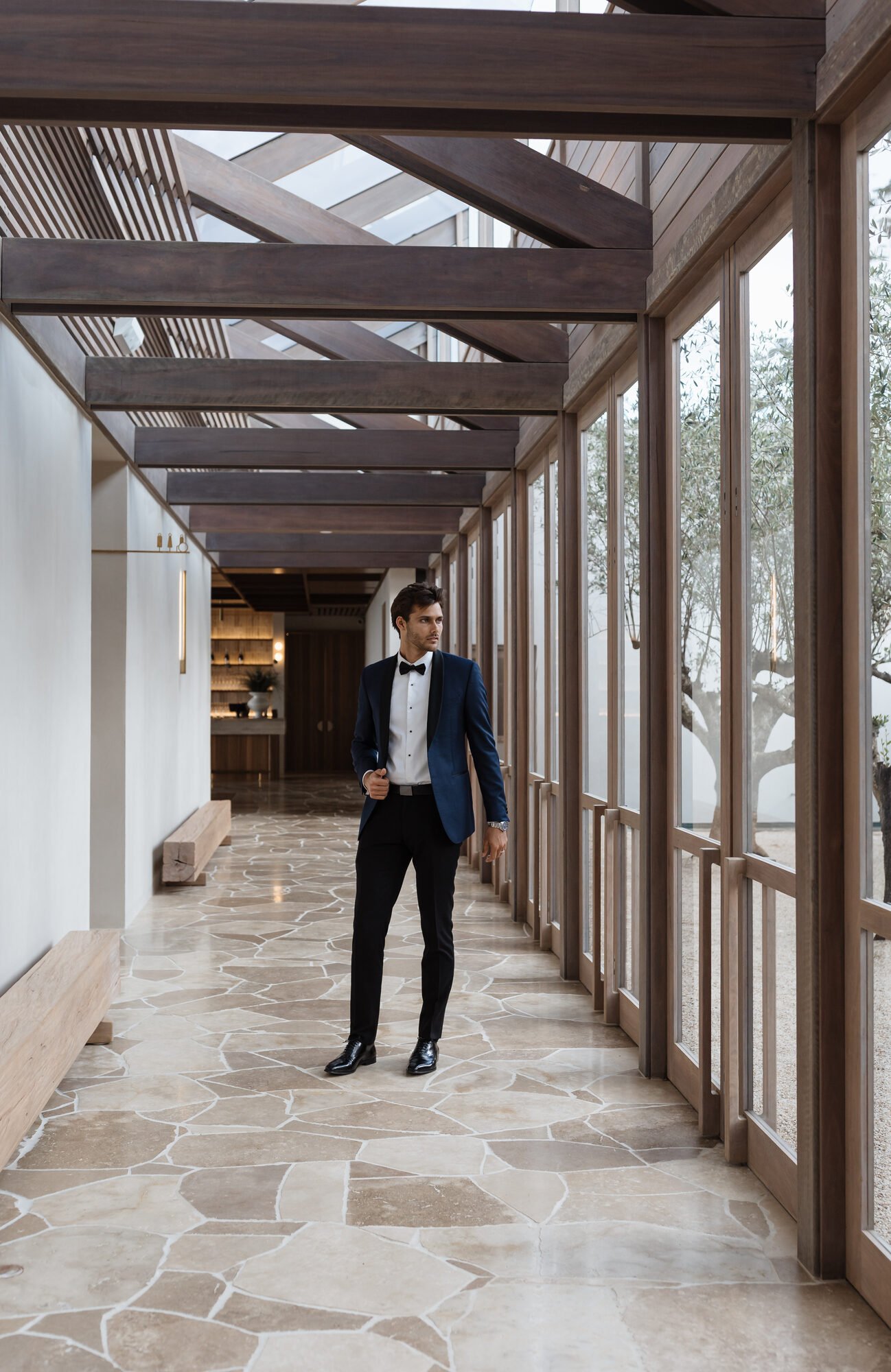 A man in a dark blue tuxedo with a black bow tie standing in a modern, sunlit interior with large floor-to-ceiling windows and patterned stone flooring.