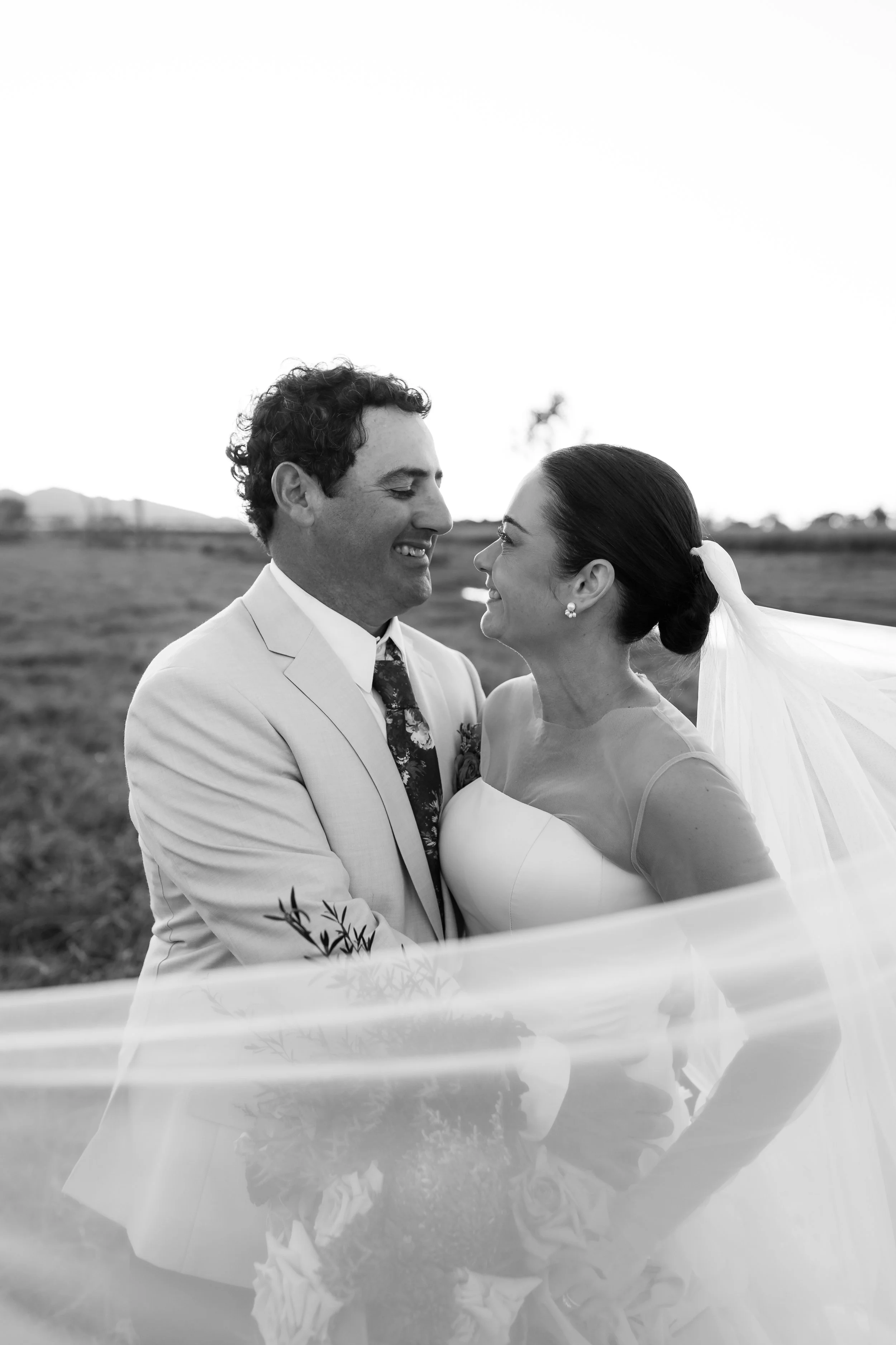 A black and white photo of a smiling man in a light-colored suit and a woman in a wedding dress looking at each other outdoors.