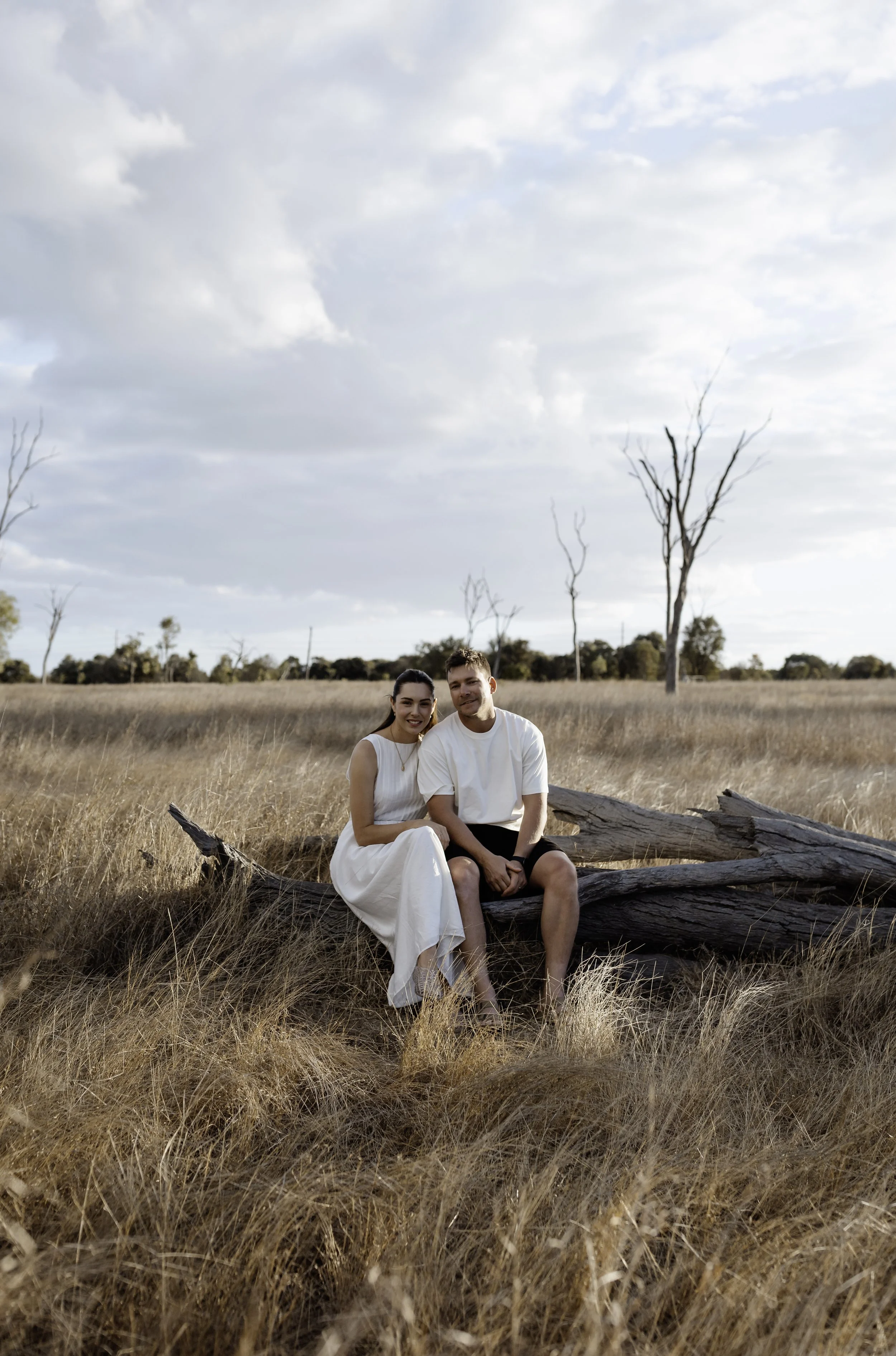 A young couple sitting on a fallen tree in a dry grassy field with a few leafless trees and cloudy sky.