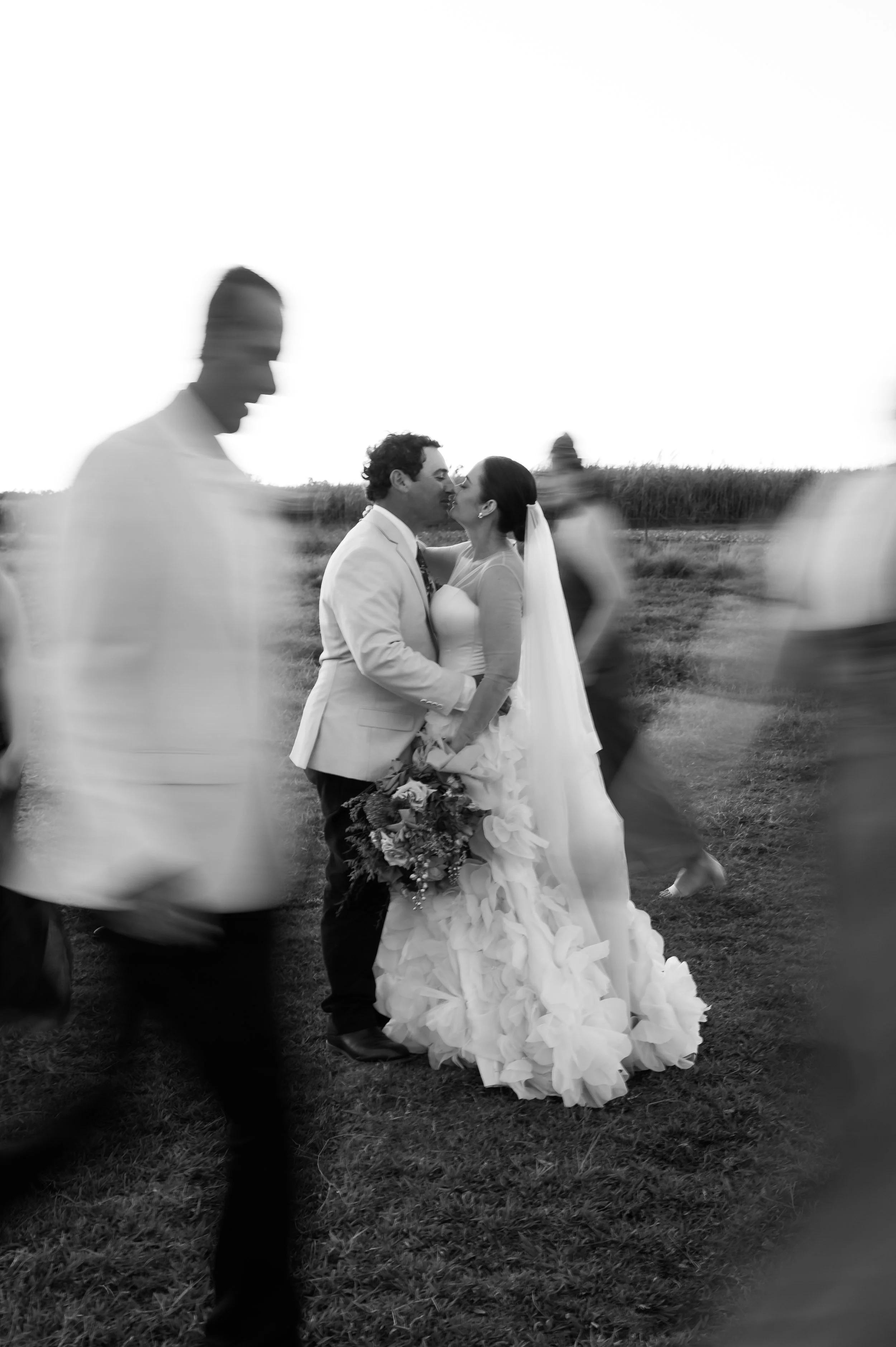 Black and white photo of a wedding with a bride and groom kissing surrounded by blurred people, outdoors in a field.