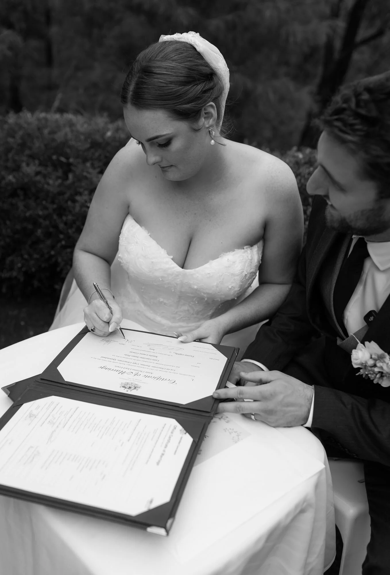 A black and white photograph of a woman in a wedding dress signing a marriage certificate, with a man next to her holding the document, outdoors with trees in the background.