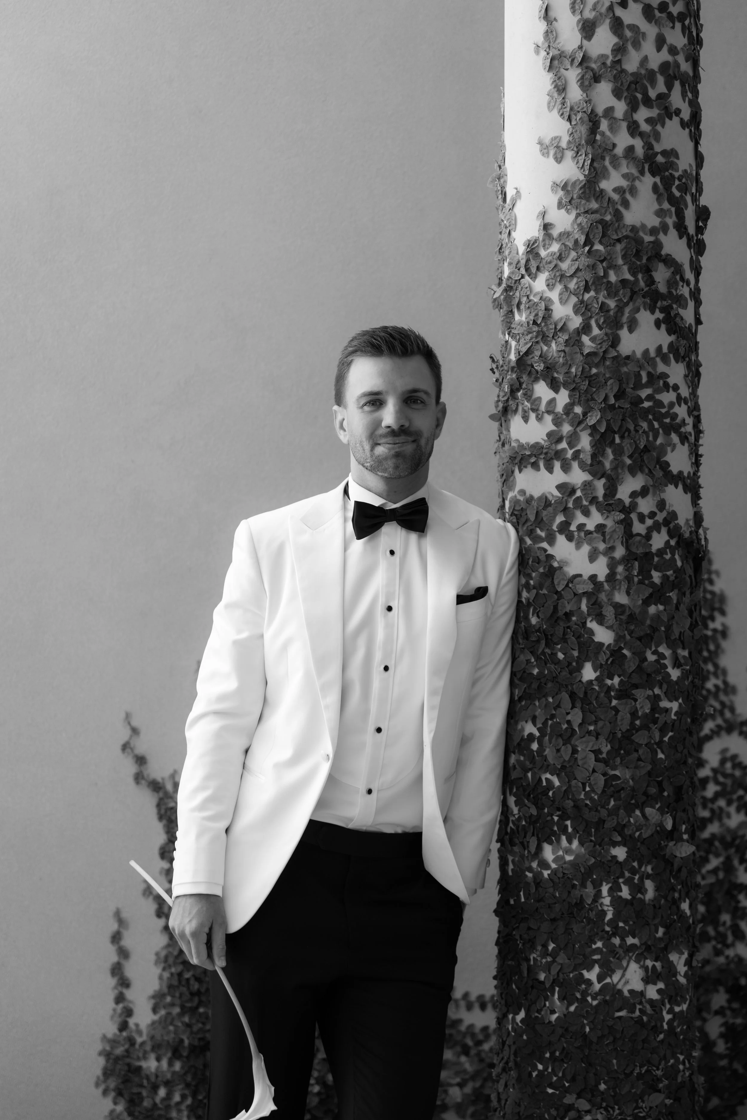 A man in a tuxedo with a bow tie standing next to a column covered with ivy, smiling in a black-and-white photograph.