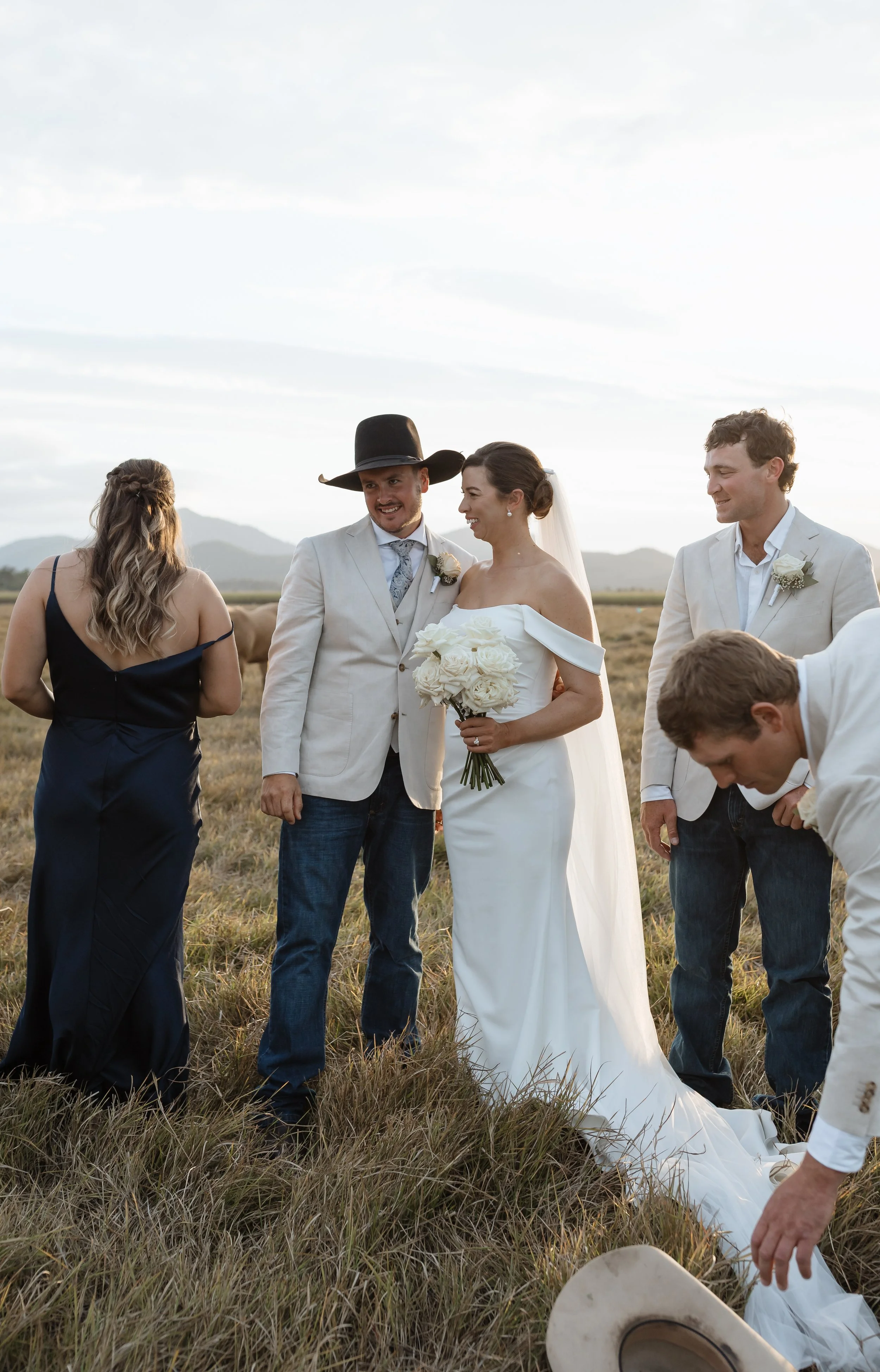 A bride holding a bouquet of white roses standing next to the groom, dressed in a light suit and cowboy hat, during an outdoor wedding ceremony in a field with mountains in the background. Wedding guests are around, dressed formally.