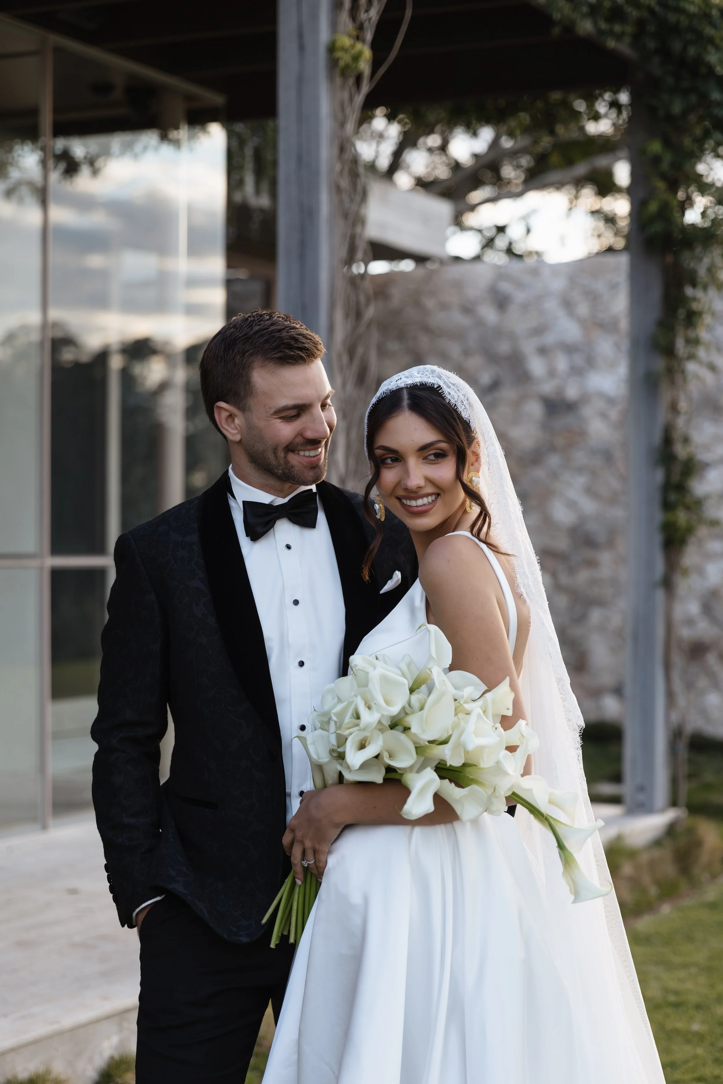 A bride and groom smiling outdoors, with the bride holding a bouquet of white calla lilies, and the groom in a black tuxedo with a bow tie.