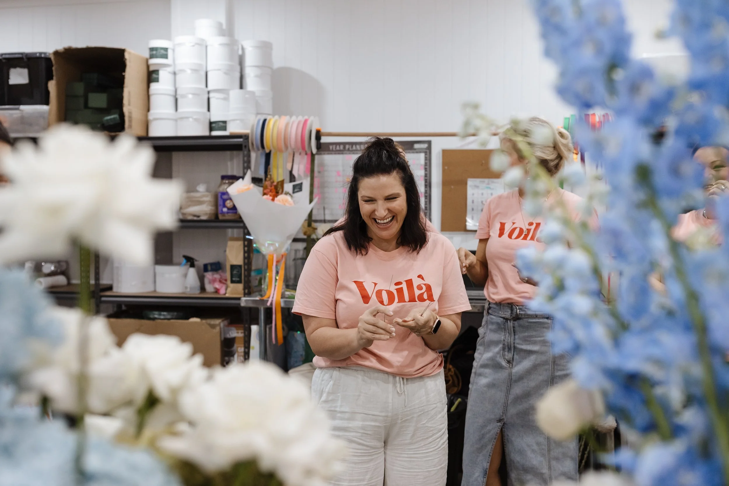 Two women in a flower shop, one smiling and looking down, the other partially visible, surrounded by flowers and floral supplies.
