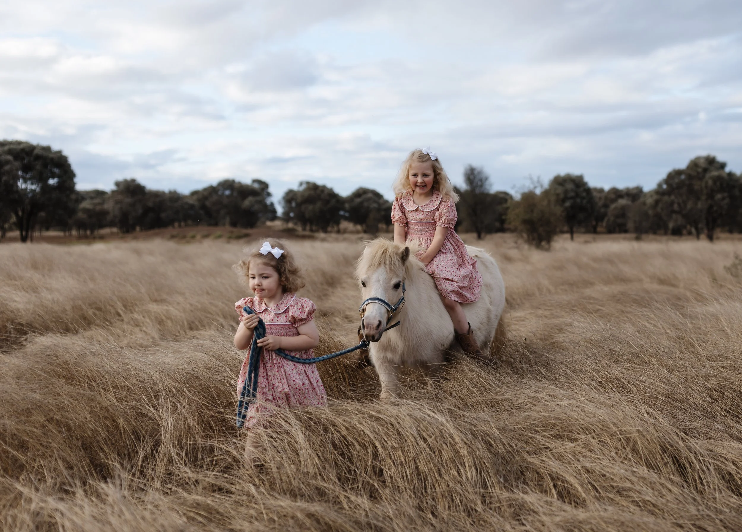 Two young girls in pink dresses riding a white pony through a grassy field with trees and cloudy sky in the background.