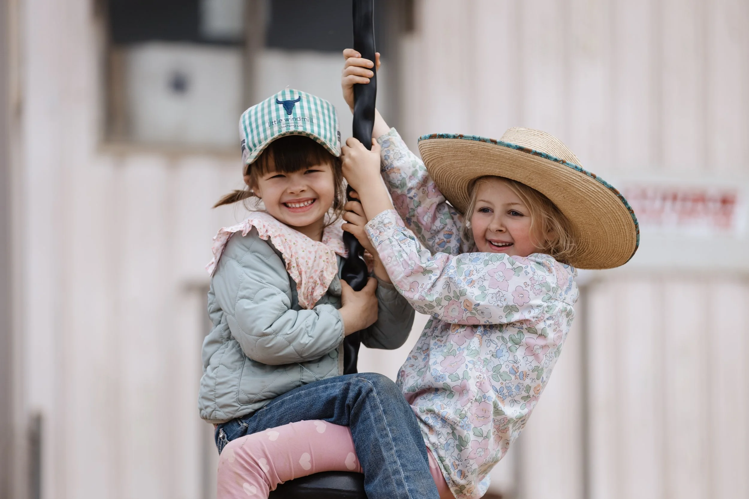 Two young girls smiling and playing together outdoors, with one girl wearing a large straw hat and the other girl wearing a checkered cap.