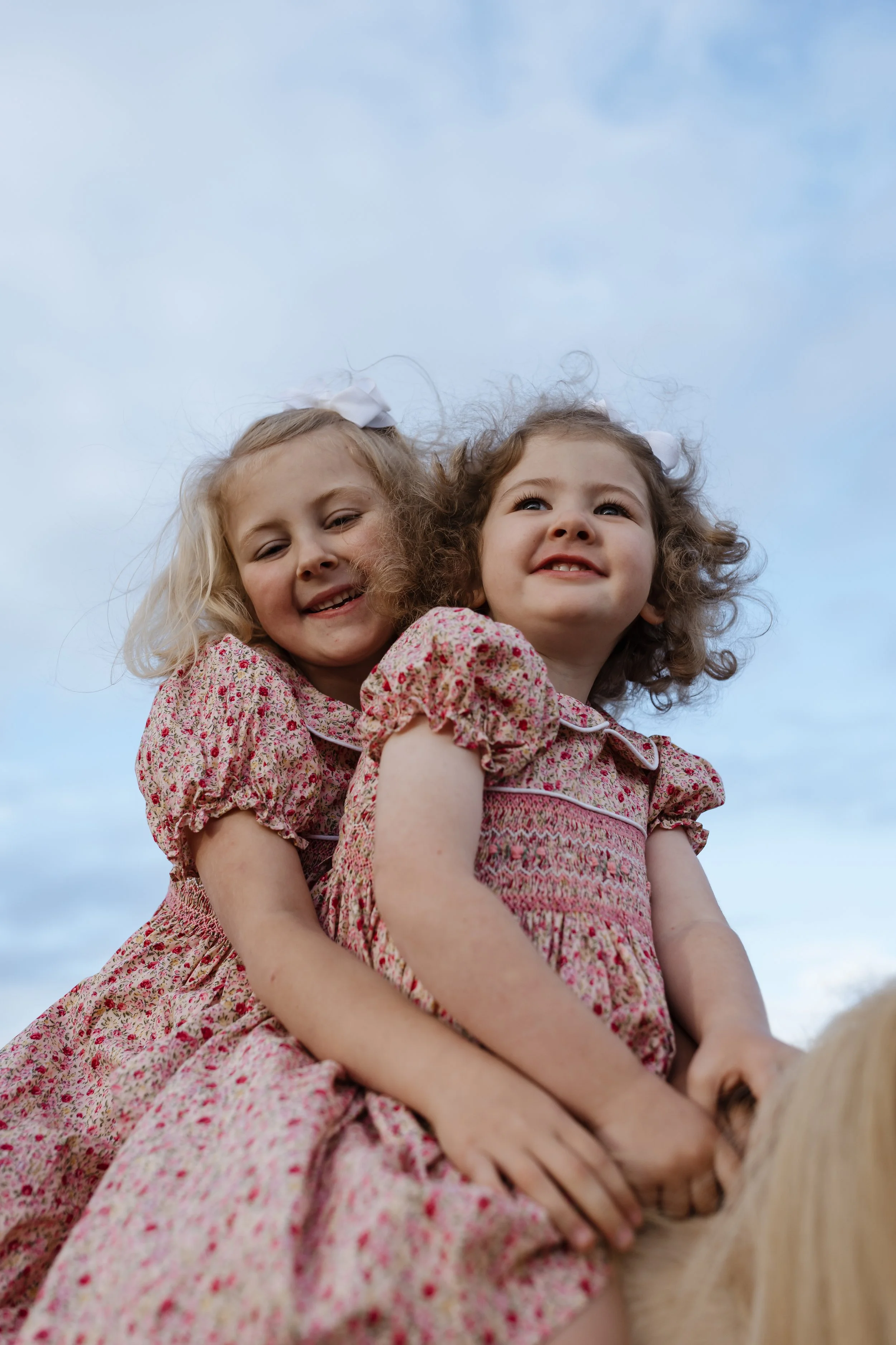 Two young girls wearing matching pink floral dresses with bows in their hair are riding a horse, with a blue sky in the background.