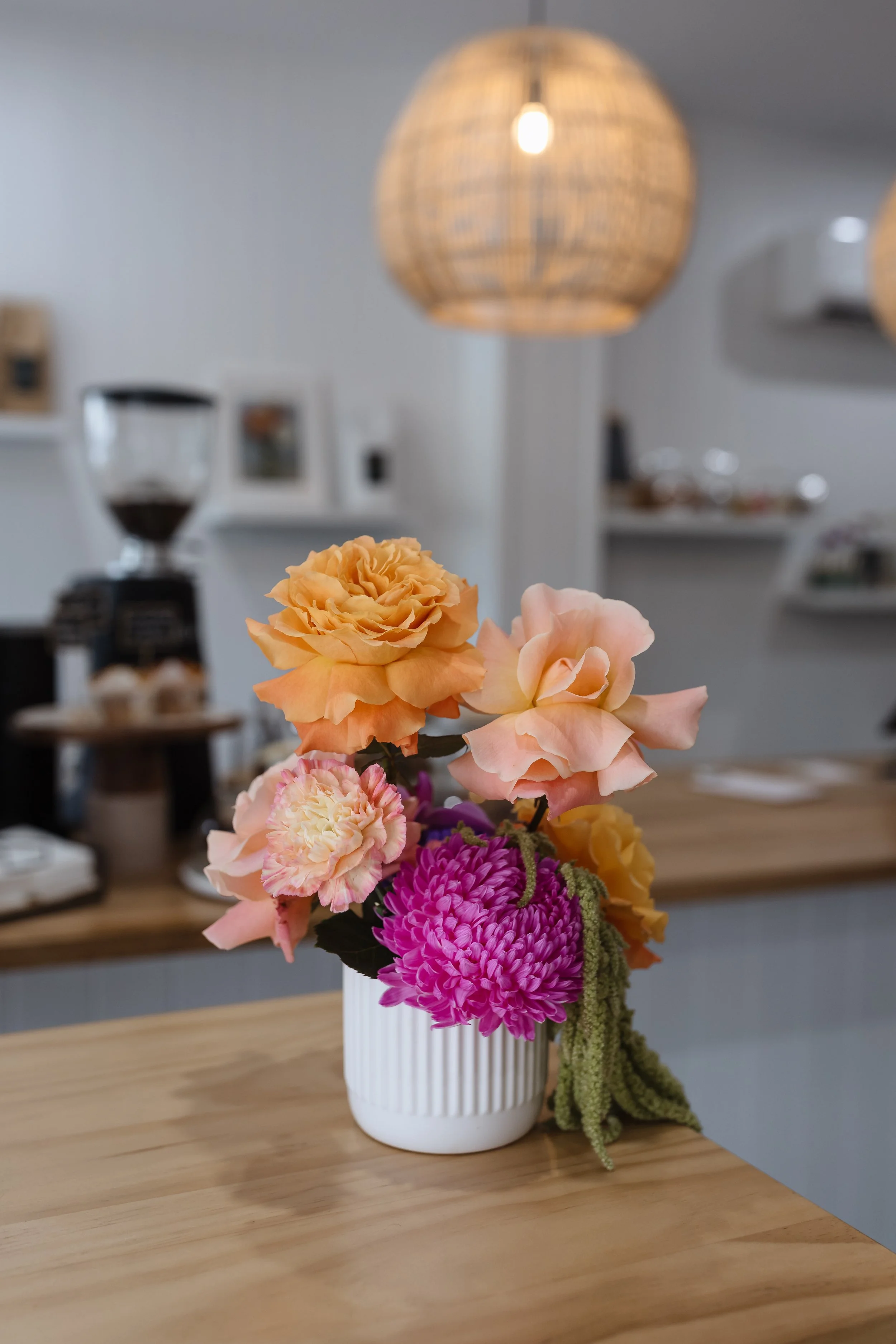 A white ribbed vase containing a colorful arrangement of artificial flowers, including peach, pink, and purple blooms, placed on a wooden table in a cozy room.