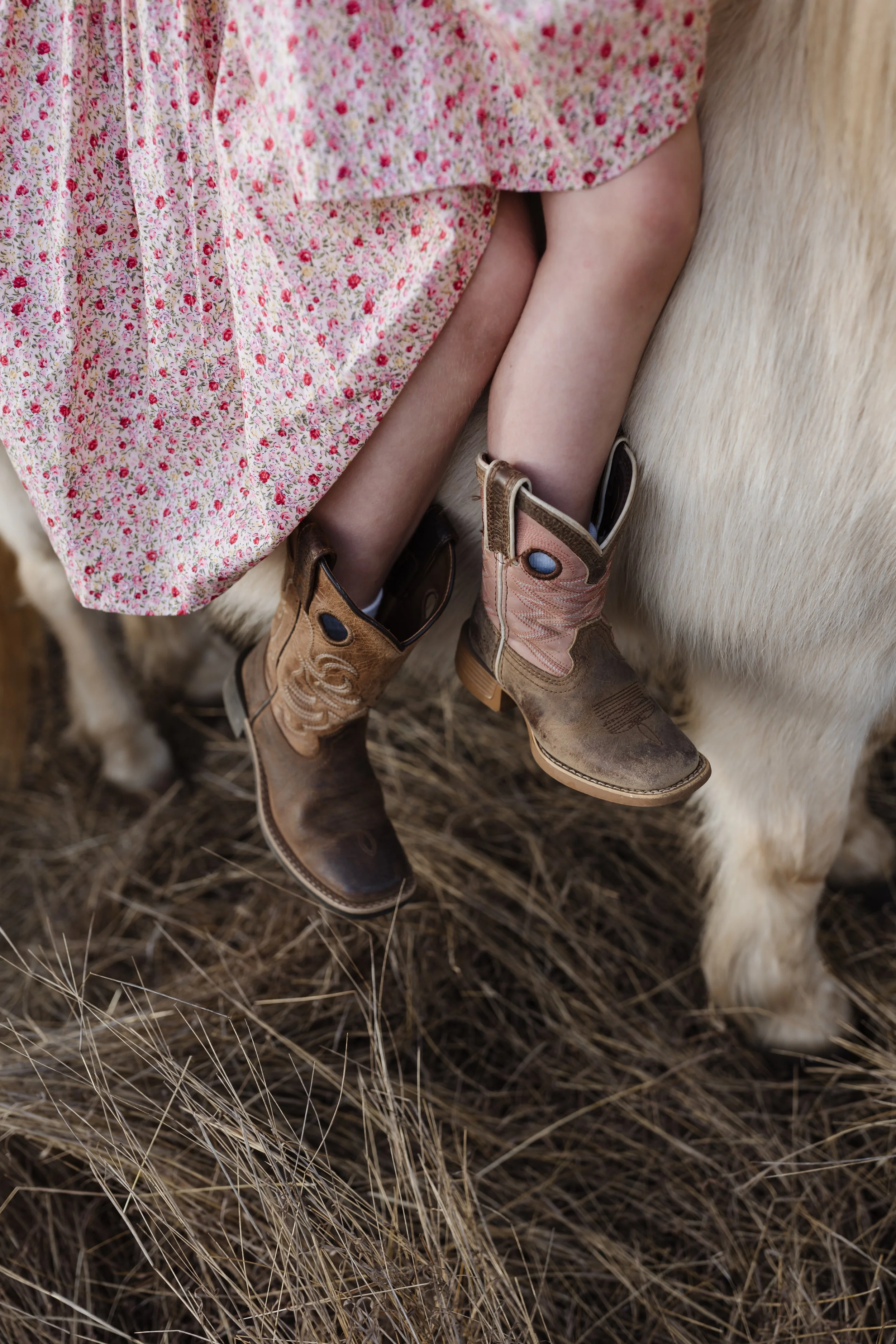 Close-up of a child's legs and feet wearing cowboy boots, sitting on a cowhide rug, with a pink floral dress visible.