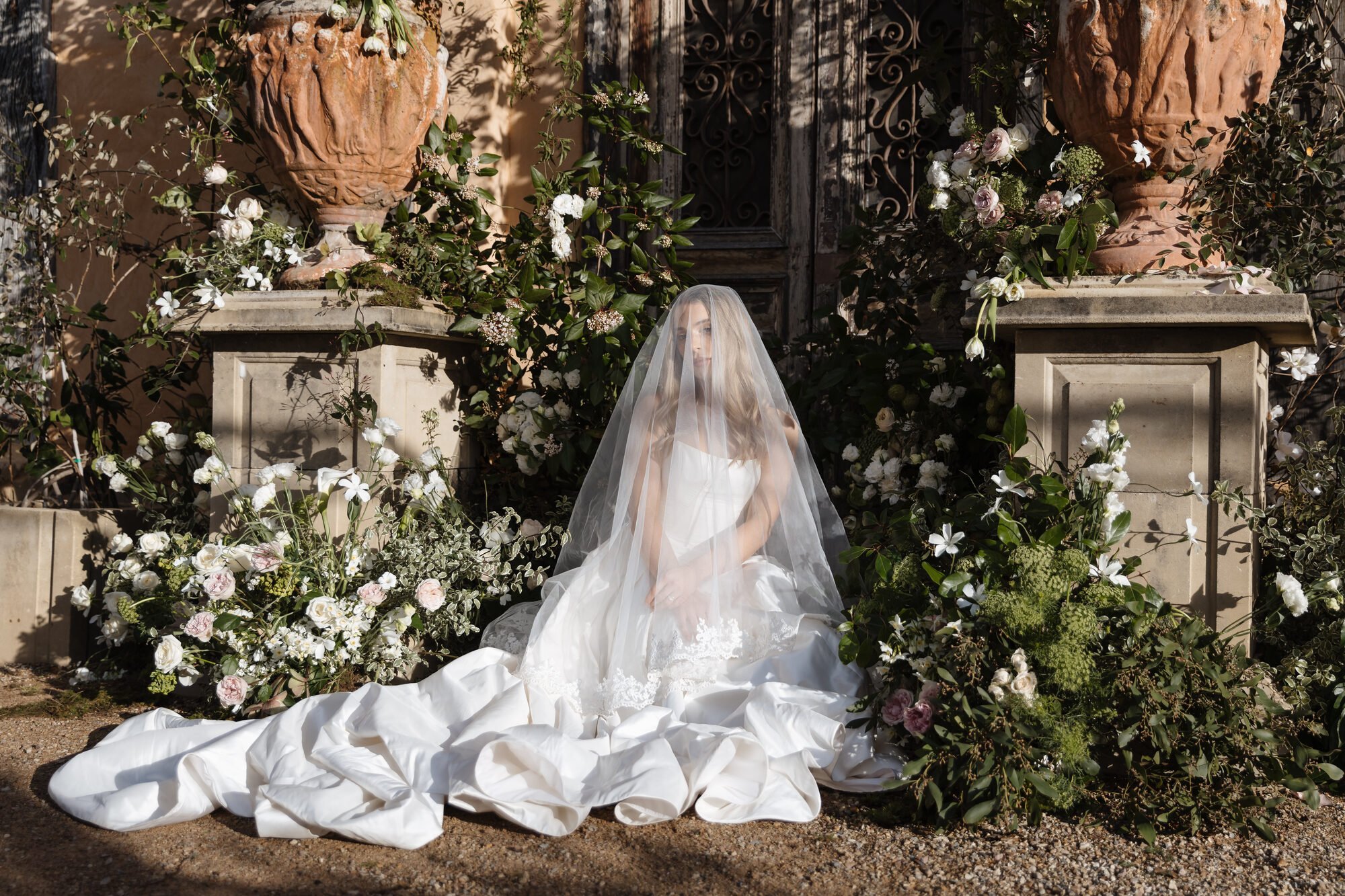 A woman dressed in a white wedding gown and veil sitting outdoors in front of large decorative urns filled with flowers, on a stone ledge, with a wrought iron gate and a beige wall behind her, surrounded by lush greenery and flowers.