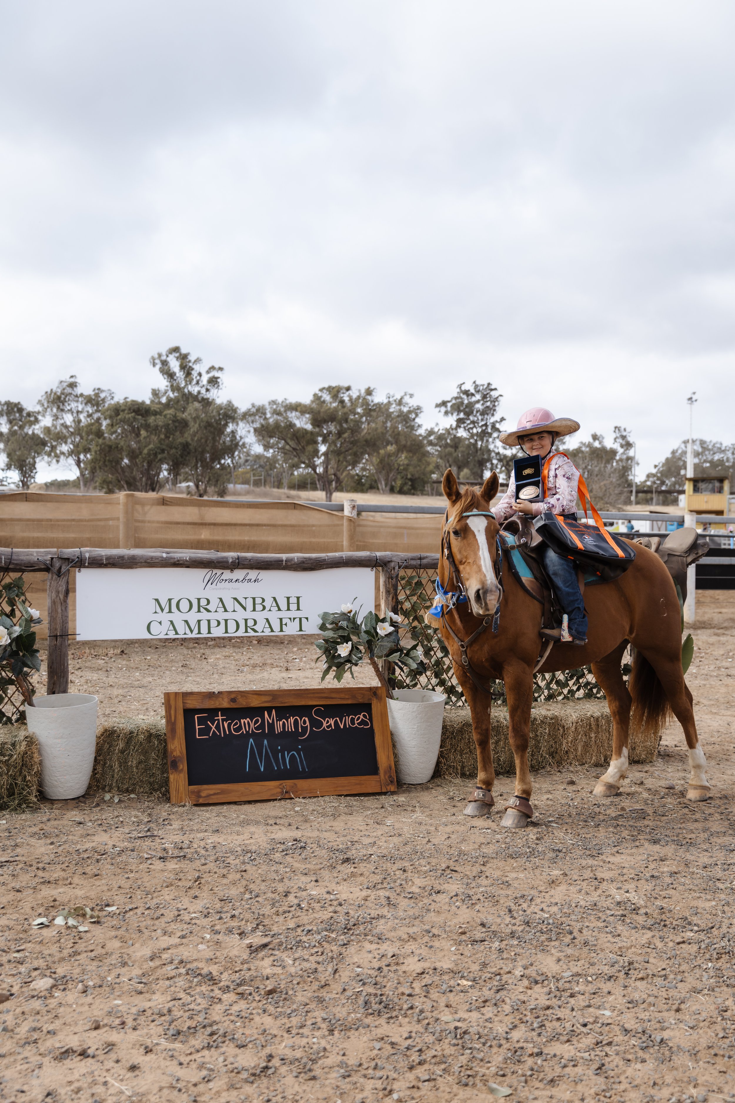 A young girl wearing a pink hat and floral shirt riding a brown horse, holding a box and smiling. The scene is at Morambah Campdraft, with event signs and decorations, trees in the background, and a cloudy sky.